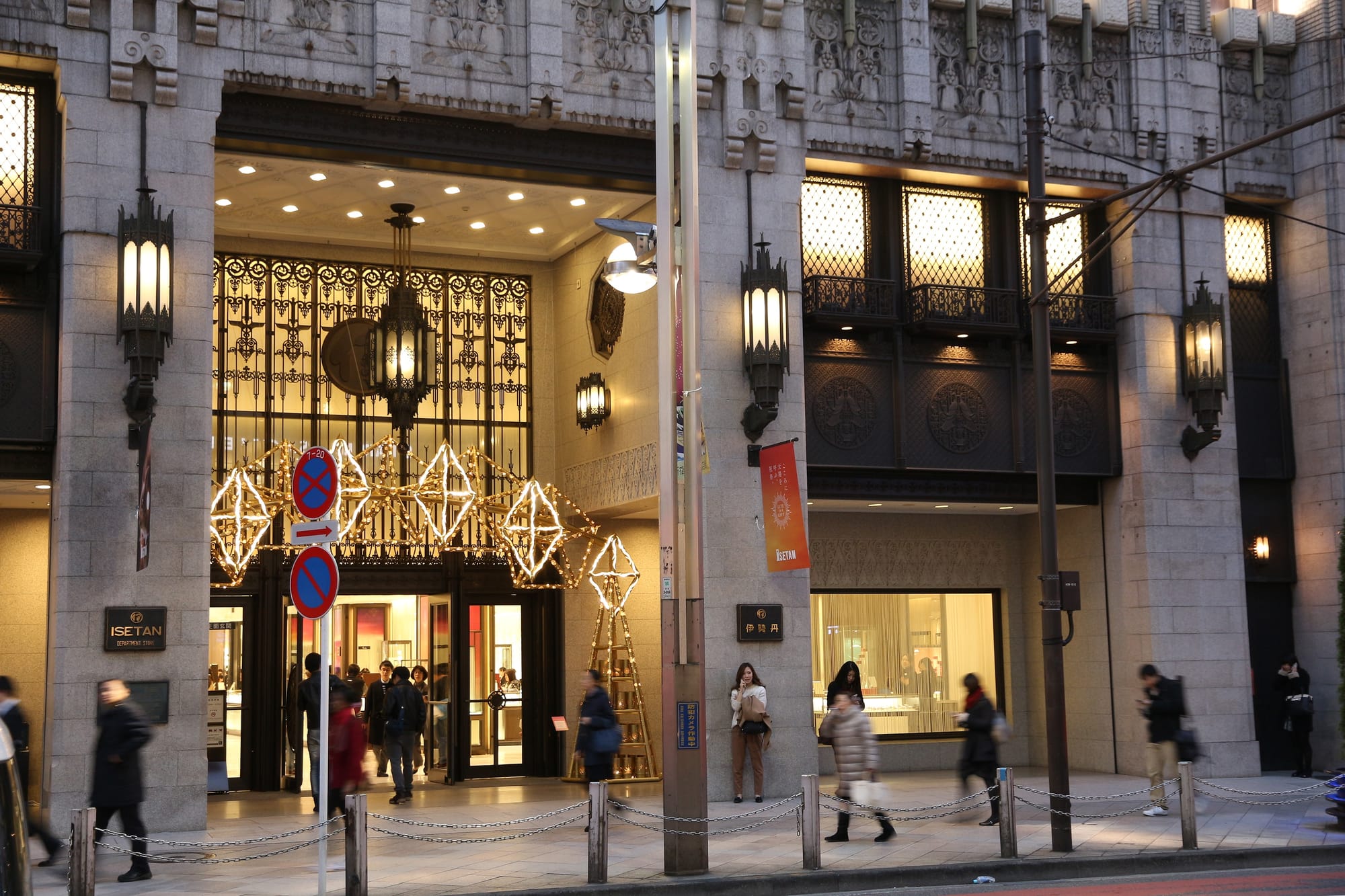 Shoppers walk by a lit entrance of a decorated building with ornate details.