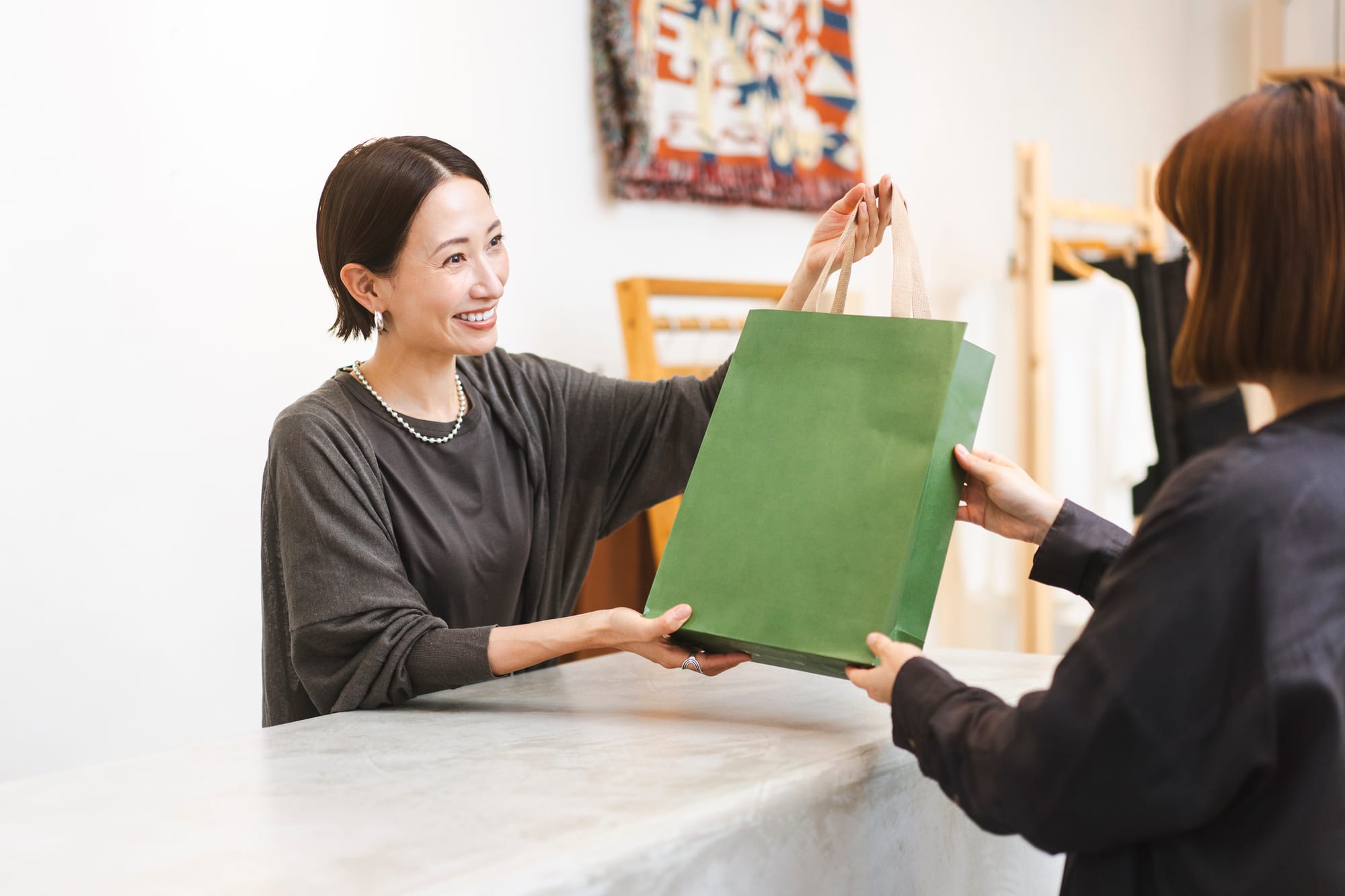 Smiling woman at a counter hands a green bag to another person.