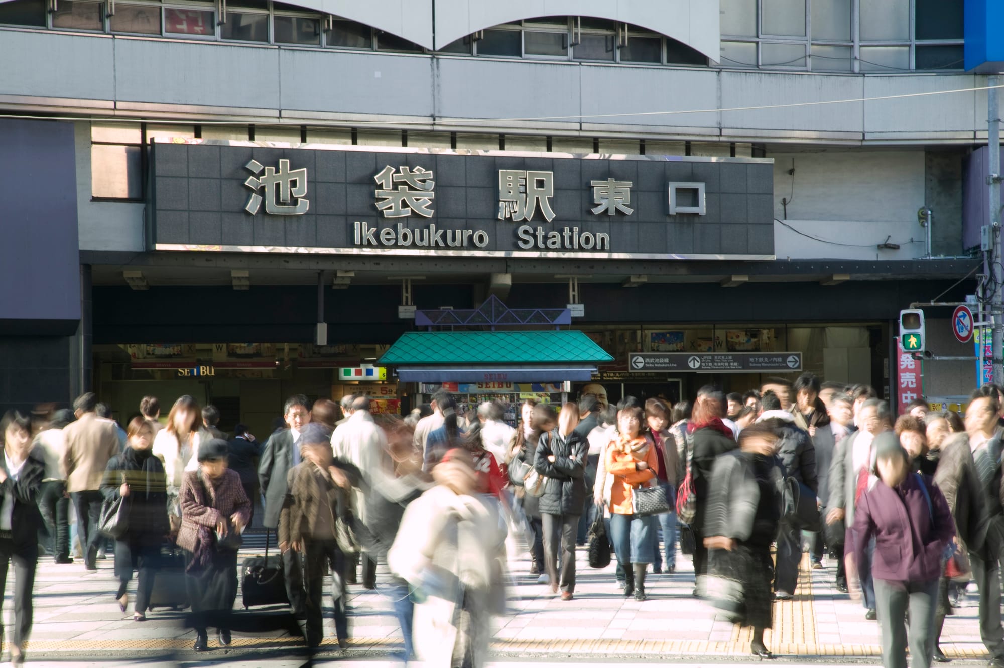 Crowd moving outside Ikebukuro Station entrance. People in winter clothes blur past.