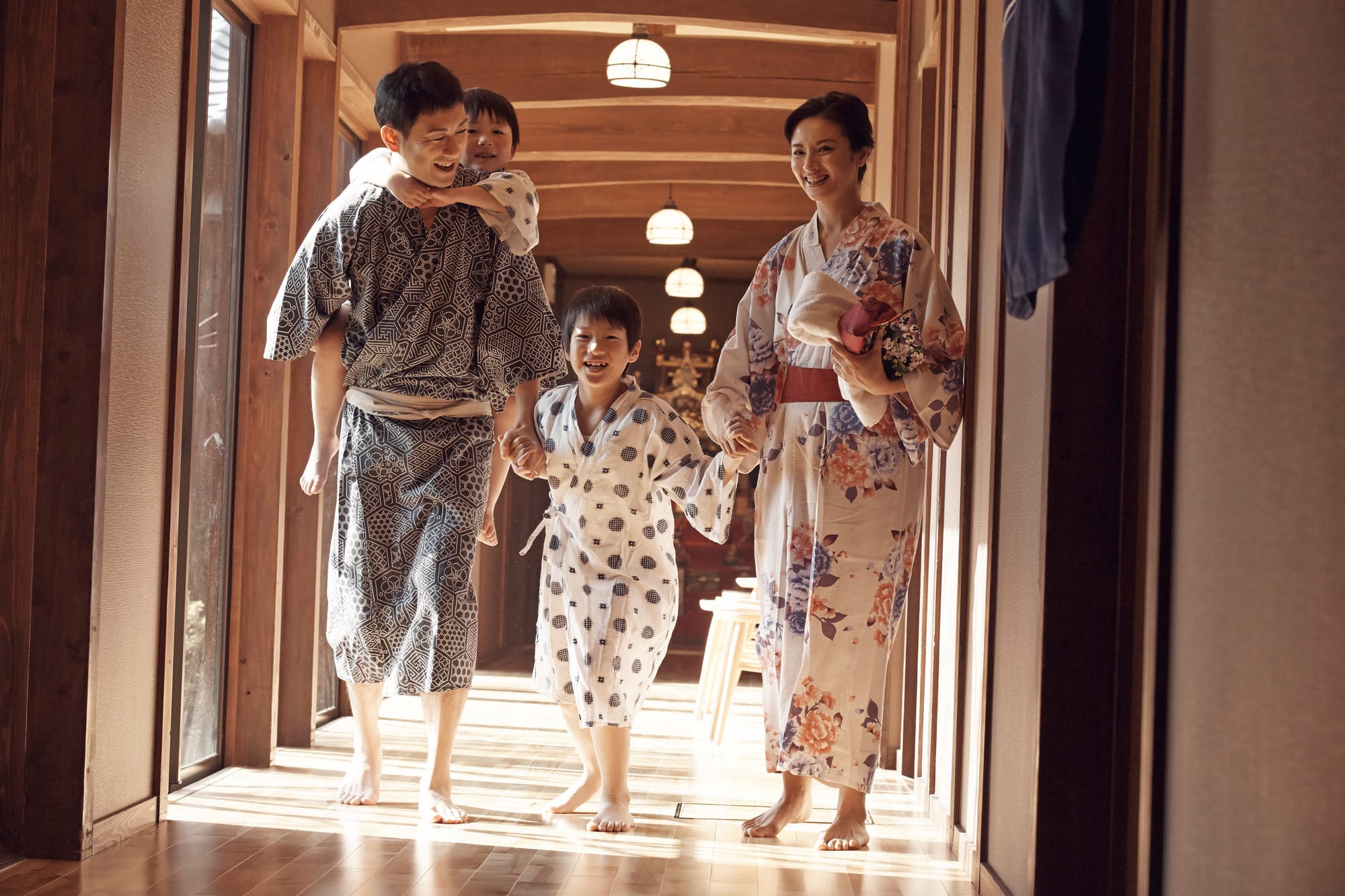 A family in patterned kimonos smiles and walks barefoot down a sunlit wooden hallway.