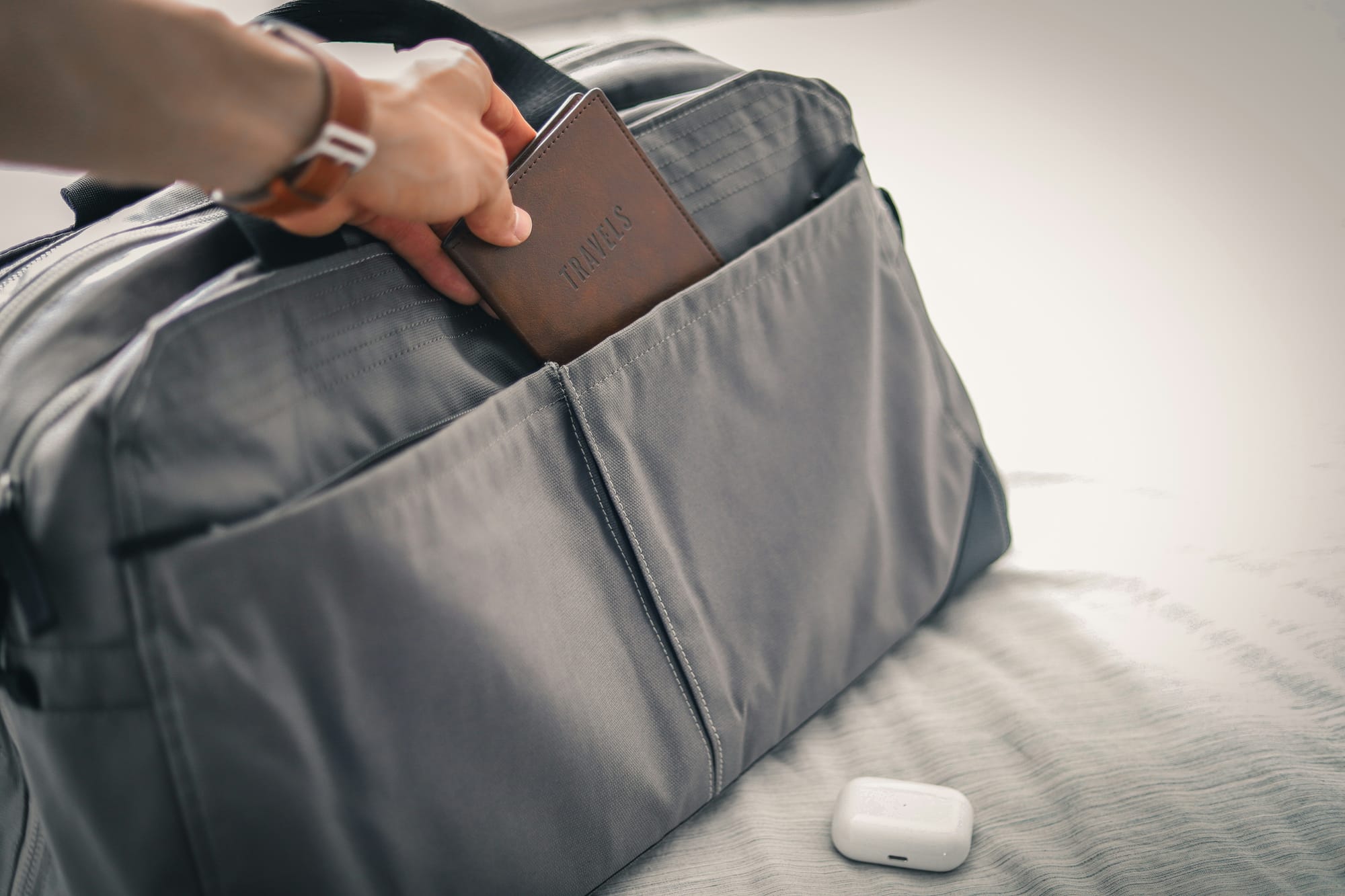 A grey travel bag sitting next to an AirPods case. A person putting a passport in the side pocket of the bag.