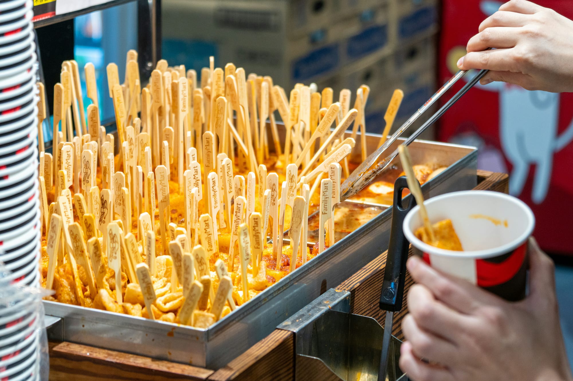 Hands using tongs to place oden from a tray into a paper cup in a Family Mart.