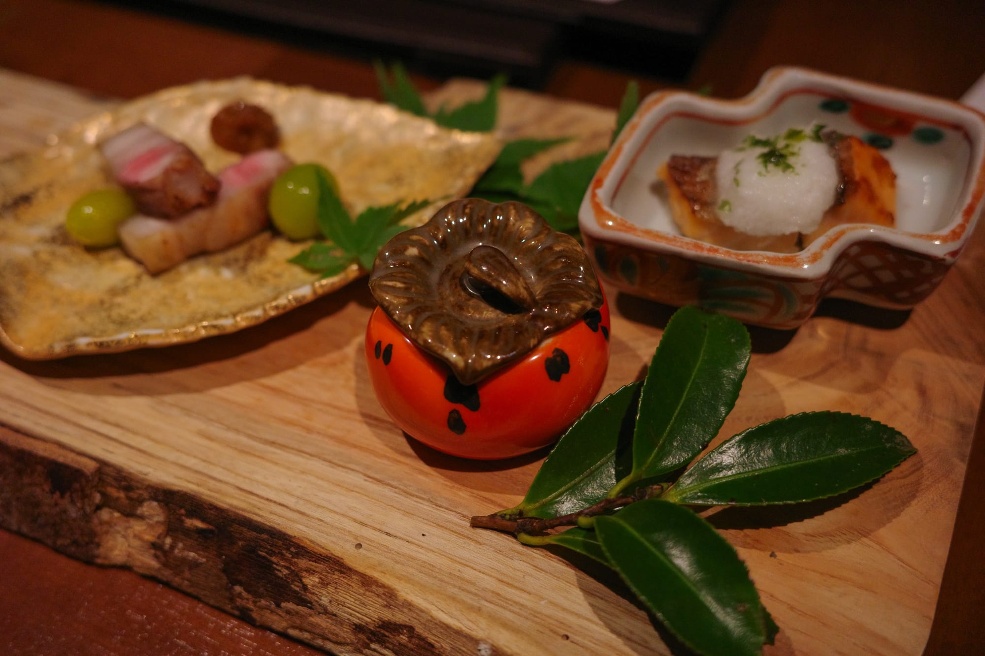 Assorted Japanese dishes on wooden board: grilled meat, gingko nuts, ceramic pumpkin, and fish with grated radish.