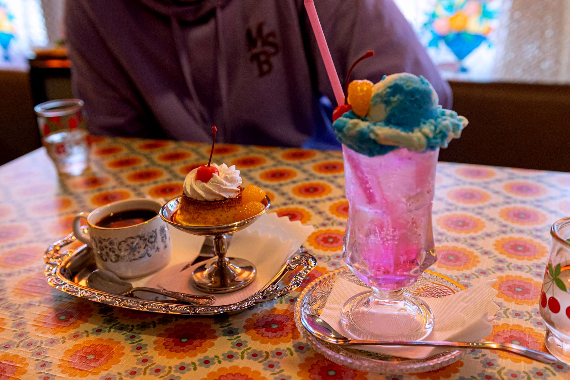Cup of coffee, flan with cherry, pink soda with blue ice cream. Vibrant patterned tablecloth.
