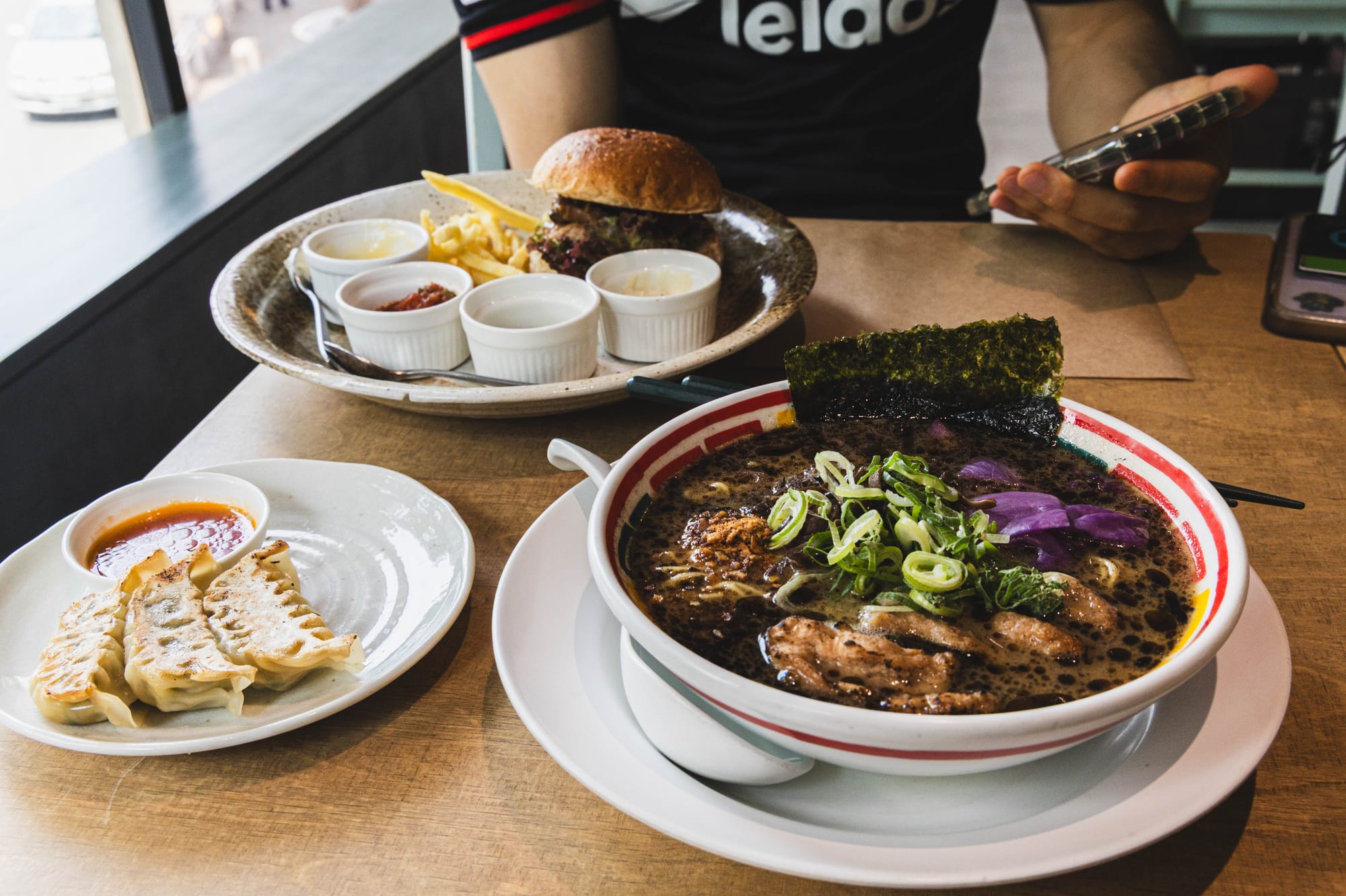 Vegan food, including ramen with toppings, gyoza with sauce, and a burger with fries on a table.