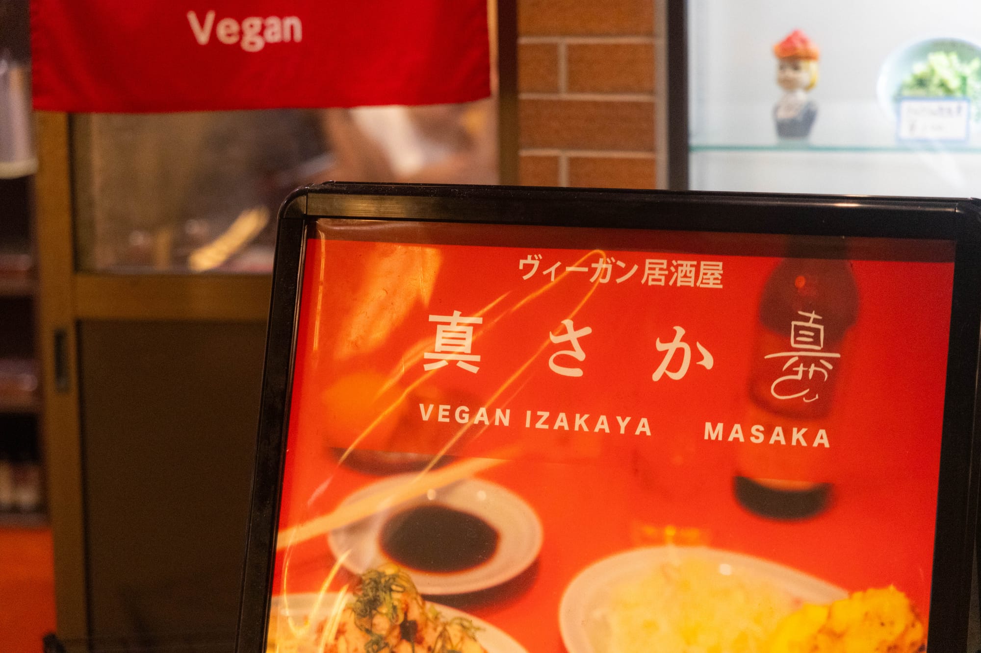 Red sign for "Vegan Izakaya Masaka" with Japanese text, set in a cozy restaurant with a brick wall and shelf display in the background.