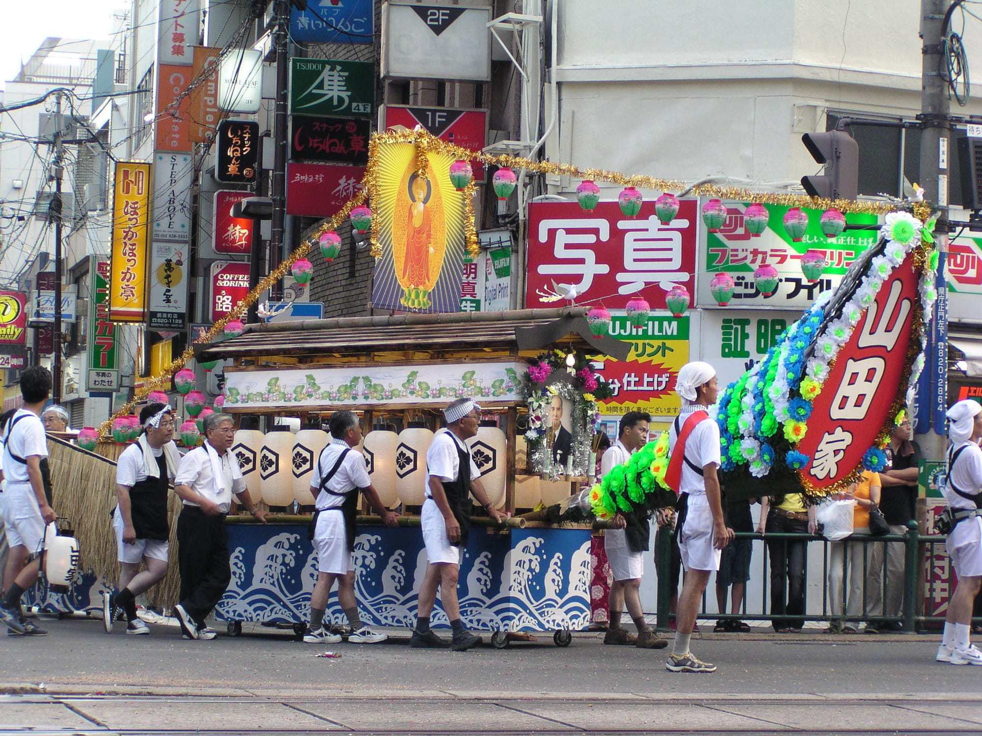 Men in traditional festival attire transport a "spirit boat" around Nagasaki for its Obon festival: the Shoro Nagashi parade. As the name implies, the spirit boat is a float representing a boat for deceased ancestors. It is decorated with lanterns, a portable altar for a deceased person complete with his portrait, a picture of a Buddhist icon, art representing waves at sea, and other details.