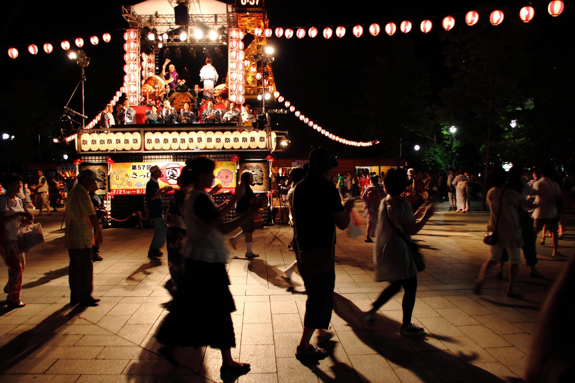 Silhouetted figures of people dancing around an illuminated yagura stage at a Bon Odori dance festival 