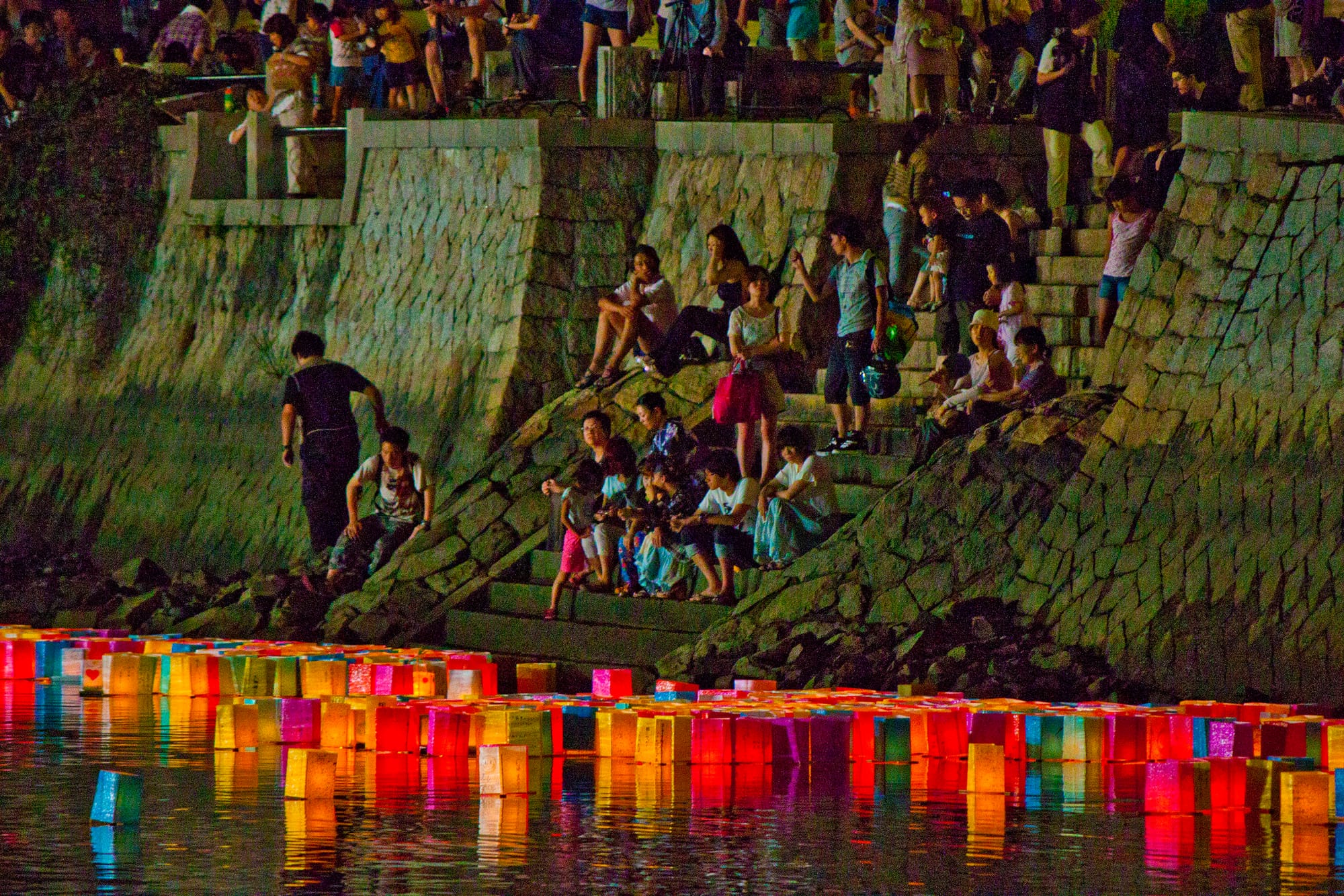 People gathering at a toro nagashi (floating lantern) ceremony in Hiroshima. Colorful lit-up lanterns float down the river.