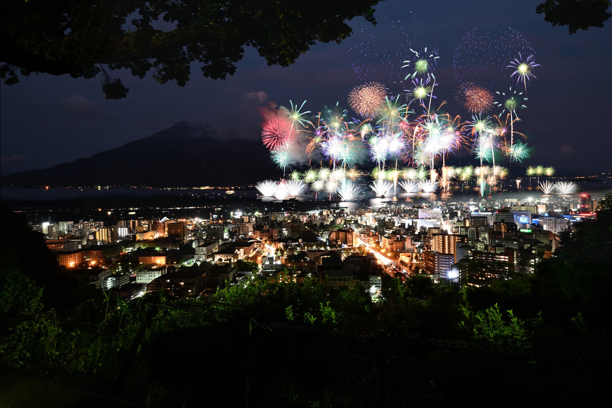 Night cityscape with vibrant fireworks lighting the sky above a brightly lit urban area. 