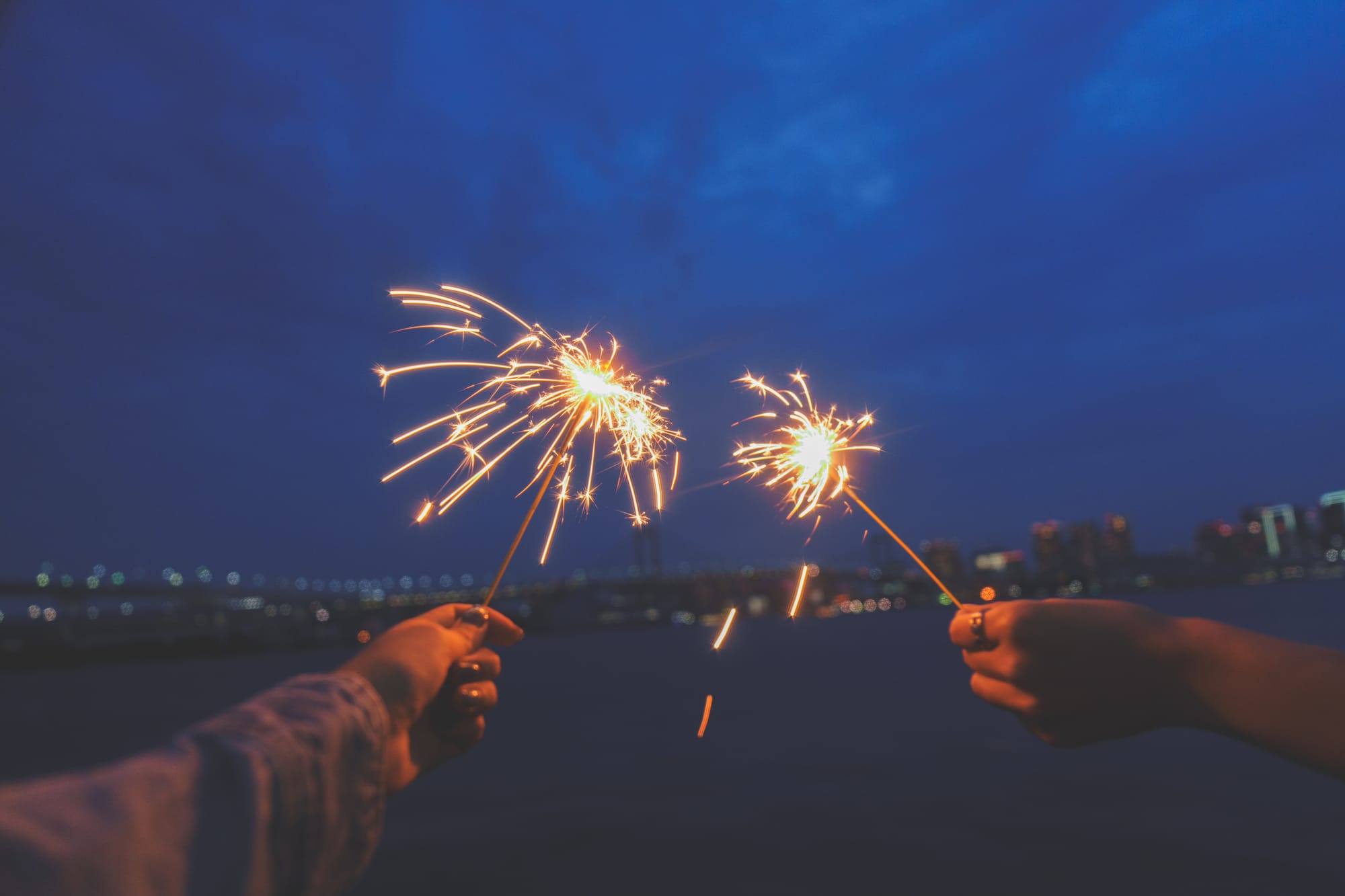 Two hands hold sparklers against a dark blue evening sky, with a cityscape and lights in the blurred background, creating a festive mood.