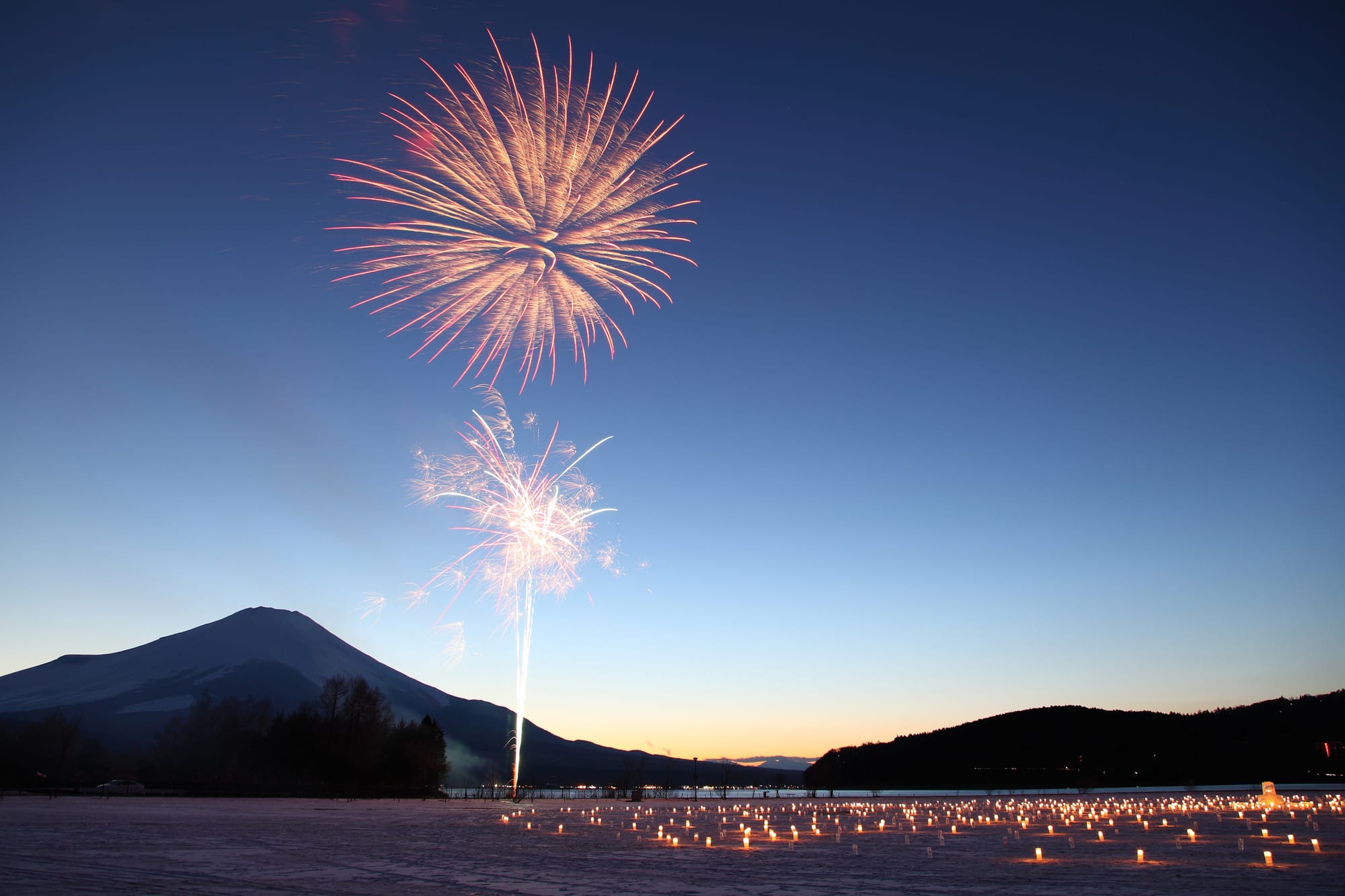 Fireworks burst over a serene landscape with Mount Fuji and a twilight sky. 
