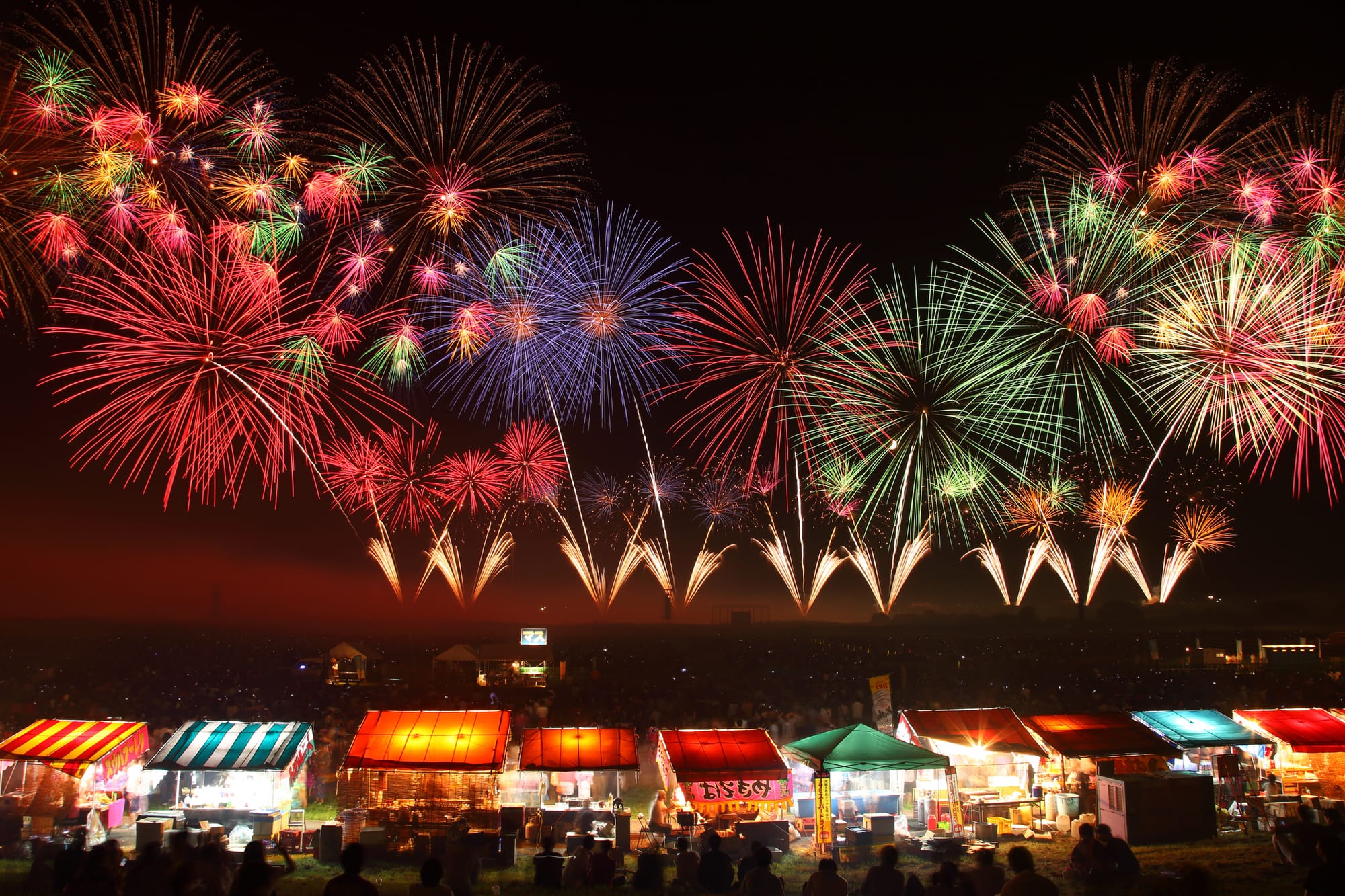 Colorful fireworks light up the night sky over vibrant market stalls with striped awnings.