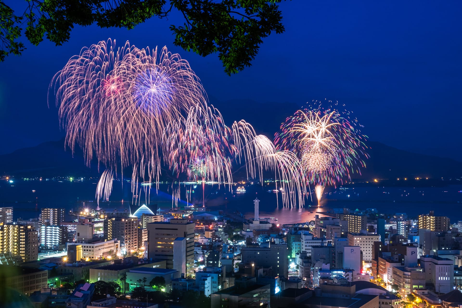 Fireworks light up the night sky above a cityscape, reflecting on the water. 