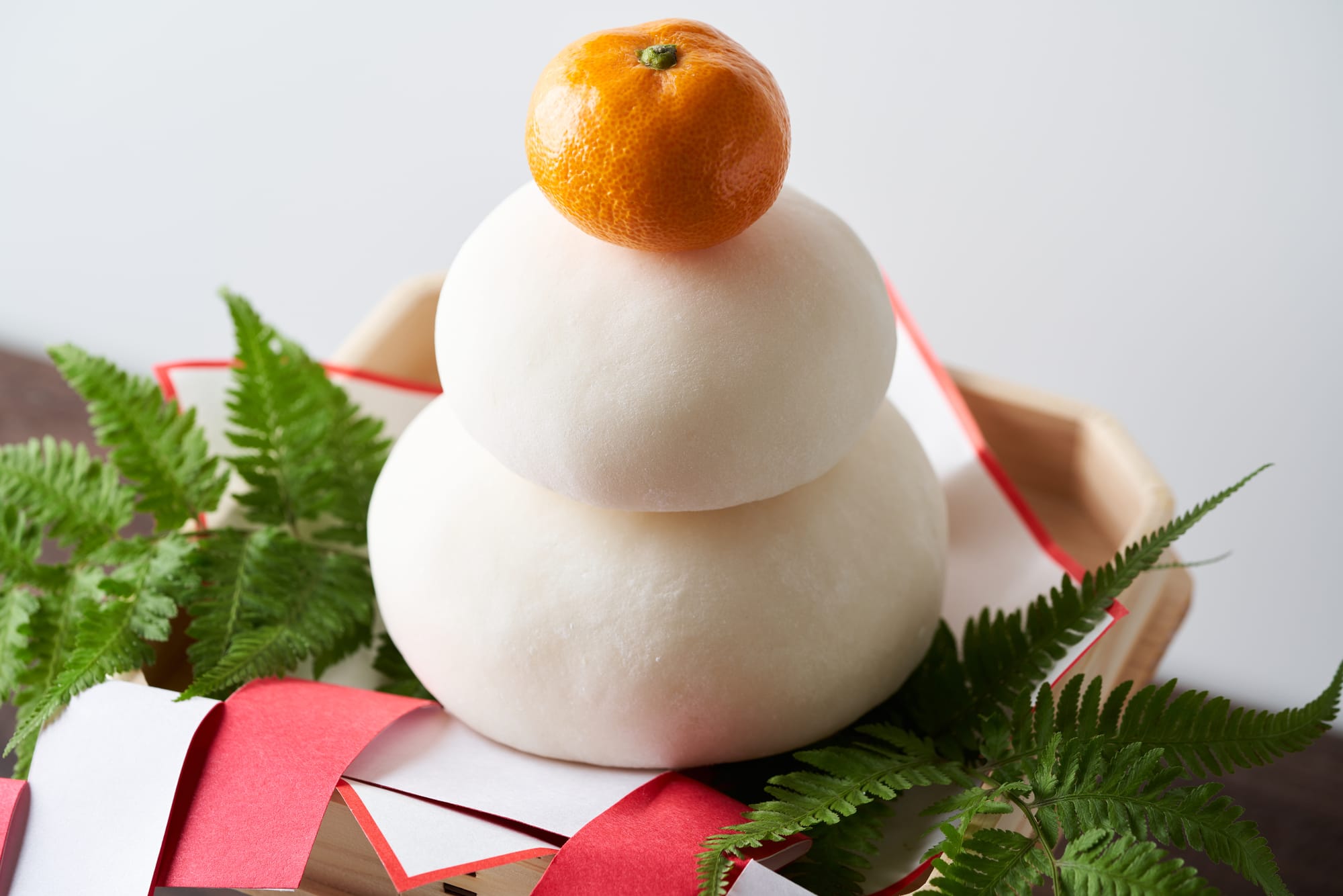 Stacked mochi with an orange on top, surrounded by green leaves and red-and-white papers.