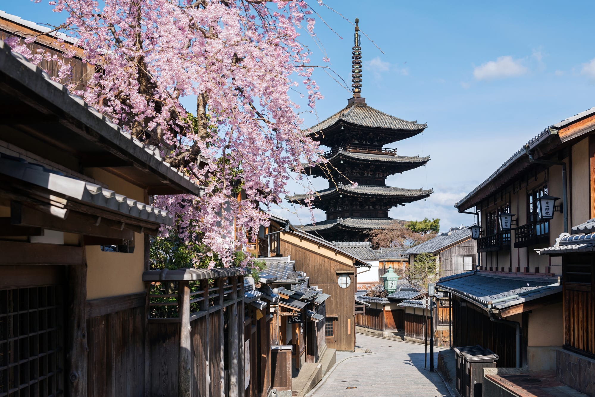 A quiet traditional street in Kyoto lined with wooden buildings and cherry blossoms, leading toward a five-story pagoda under a blue sky.