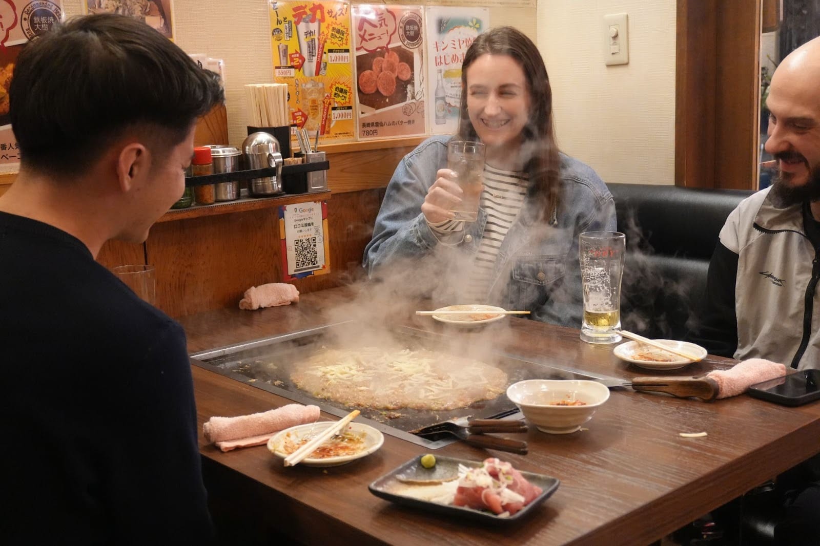 A guide and two similing guests watching their dinner, a savory pancake-like dish called monjayaki, being grilled in front of them