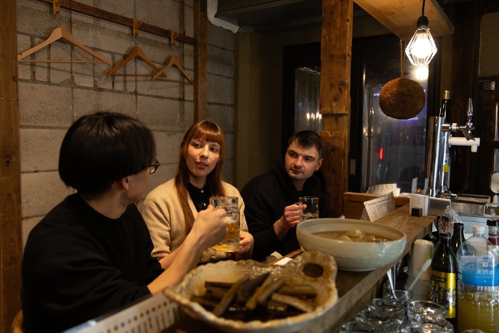 A local guide chatting with two guests from abroad at an izakaya Foreign tourists and a Japanese guide raising their mugs and glasses for a toast at an izakaya pub during one of TOMOGO!'s Japan bar-hopping tours