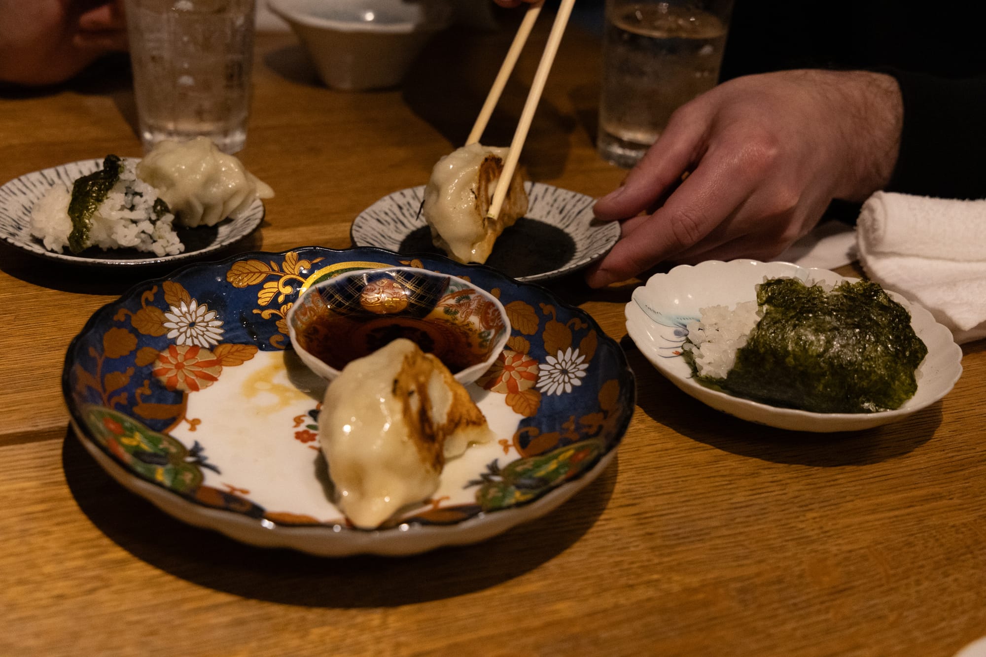 Close-up photo of snacks such as gyoza dumplings and onigiri rice balls, with a diner picking up a piece of gyoza with a chopstick and dipping it in soy sauce