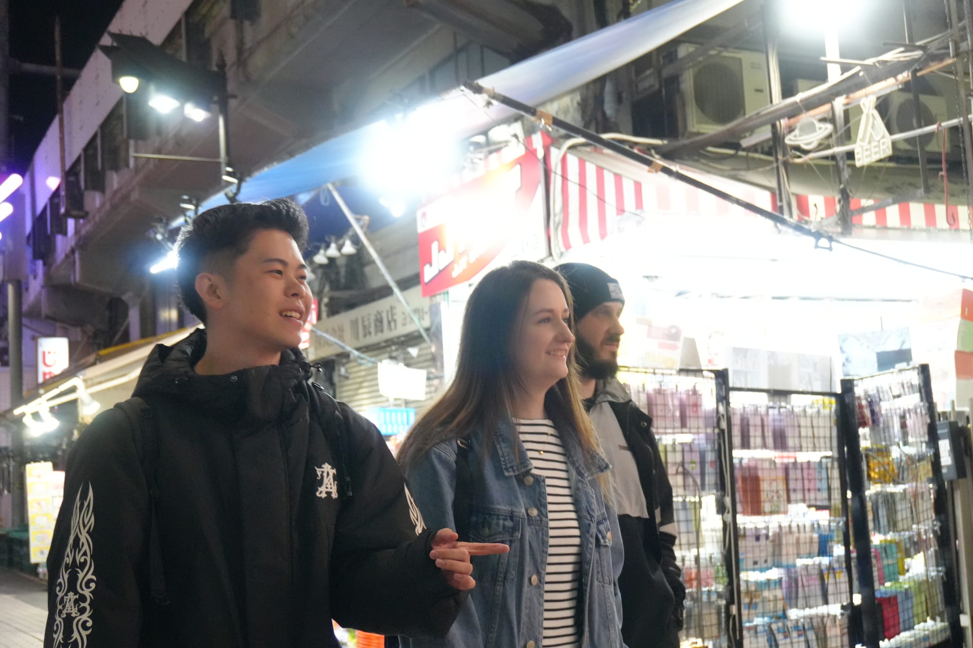 A guide pointing something out to two guests as they walk down a brightly lit alley with shops and stalls during one of TOMOGO!'s Japan bar-hopping tours