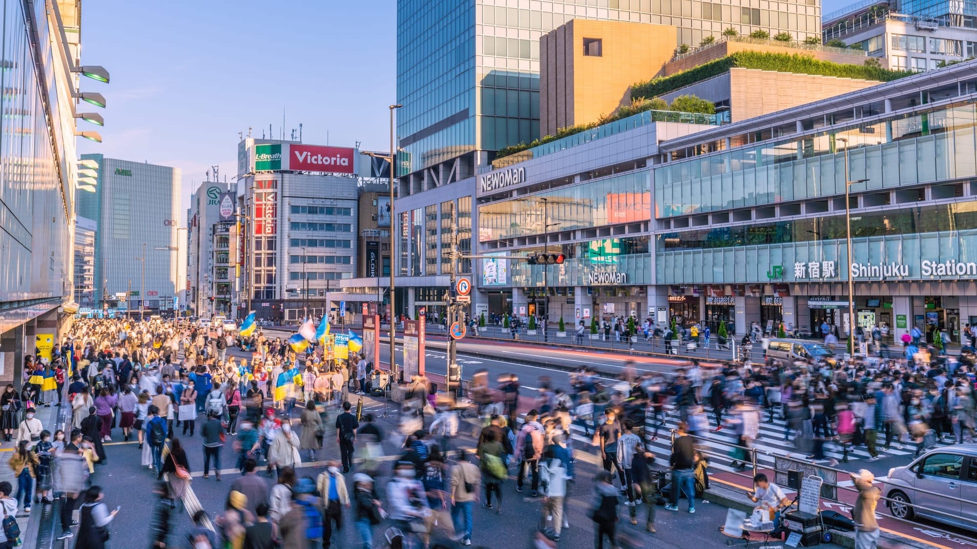 A busy street crossing outside Shinjuku Station in Tokyo, with motion-blurred crowds and the JR station entrance sign visible on the right.