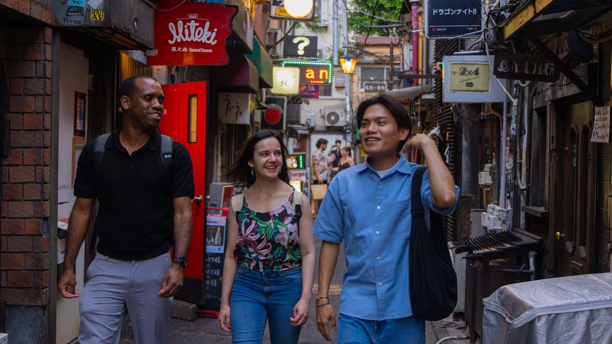 Three people walking and smiling through the narrow alleyways of Golden Gai in Shinjuku, Tokyo, surrounded by neon signs and tiny bar entrances.