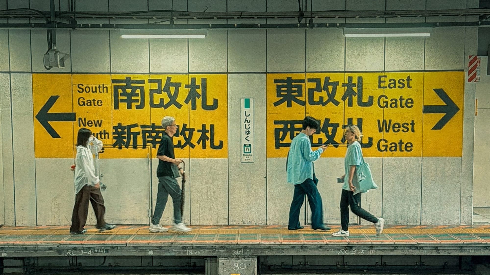 Passengers walking along a Shinjuku Station platform past yellow bilingual signs pointing to the South Gate, New South Gate, East Gate and West Gate exits.