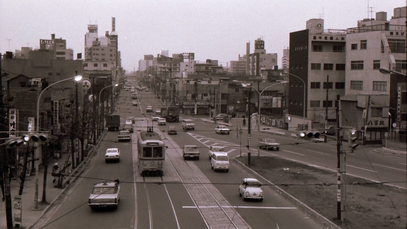 Vintage black-and-white photo of a busy Tokyo street in the 1960s, with a tram, mid-century buildings, and Japanese signage.
