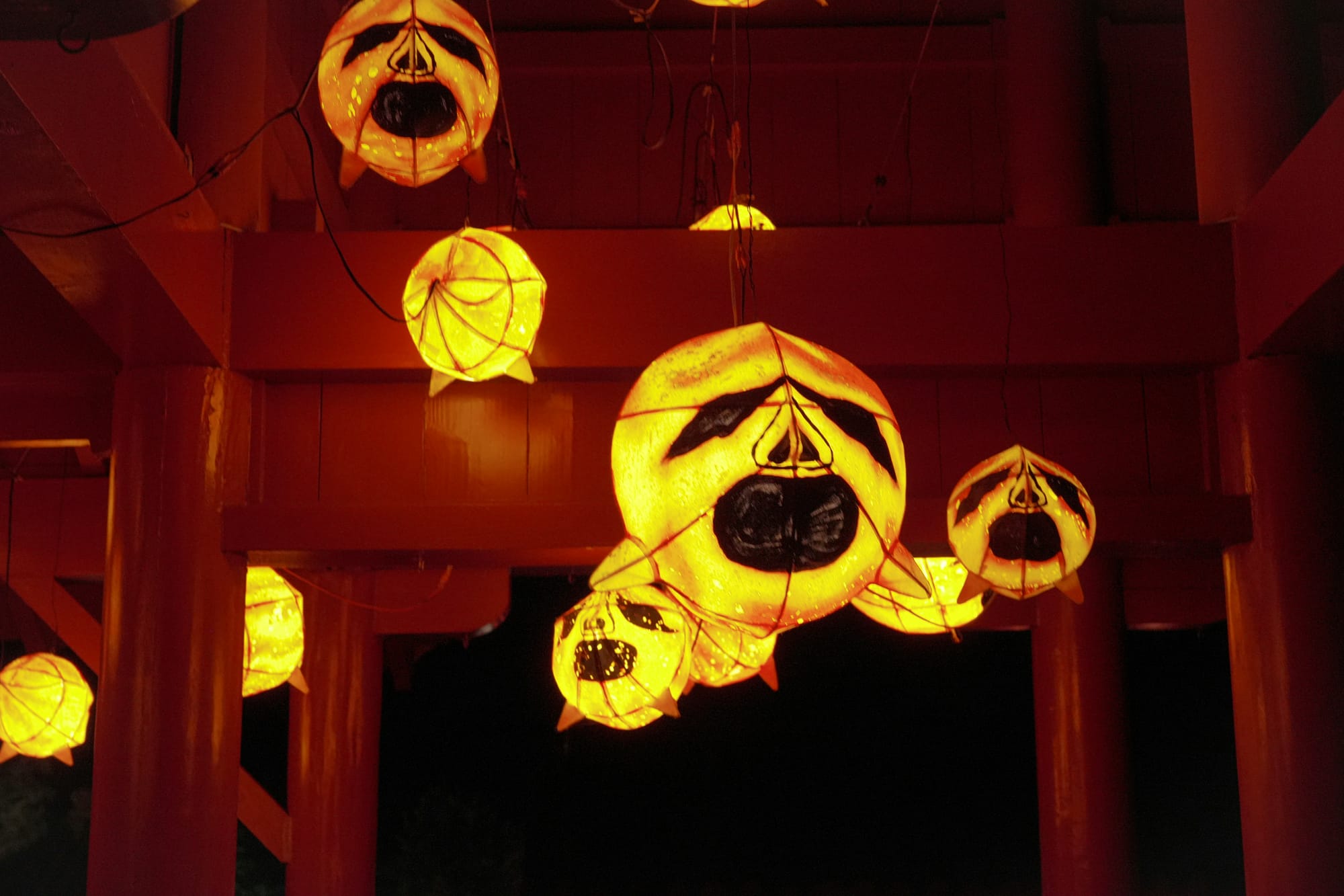 Glowing orange paper lanterns with spooky jack-o’-lantern faces hang in a dark, red-lit space, creating an eerie Halloween atmosphere.