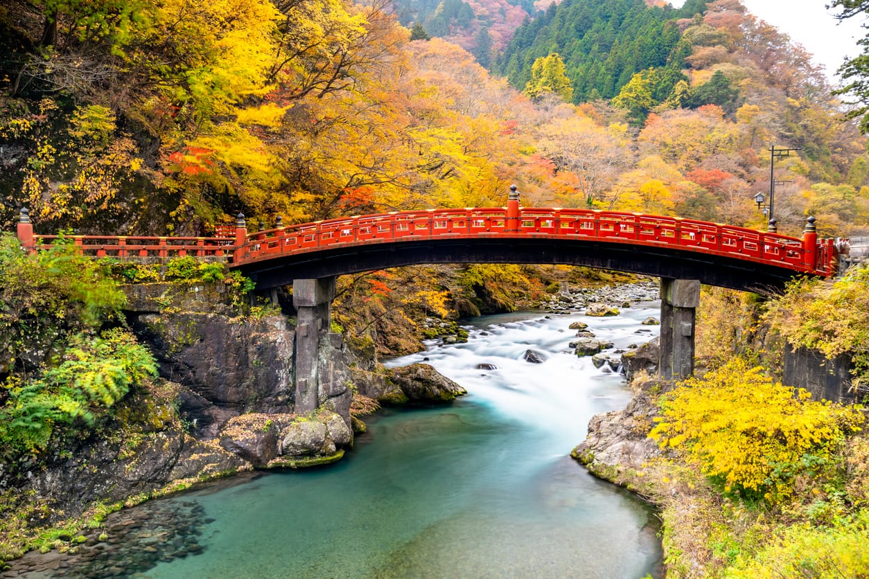 A traditional Japanese curved bridge, painted in vermillion red. The bridge crosses a river with mild rapids, and the valley is surrounded by red and gold fall foliage.