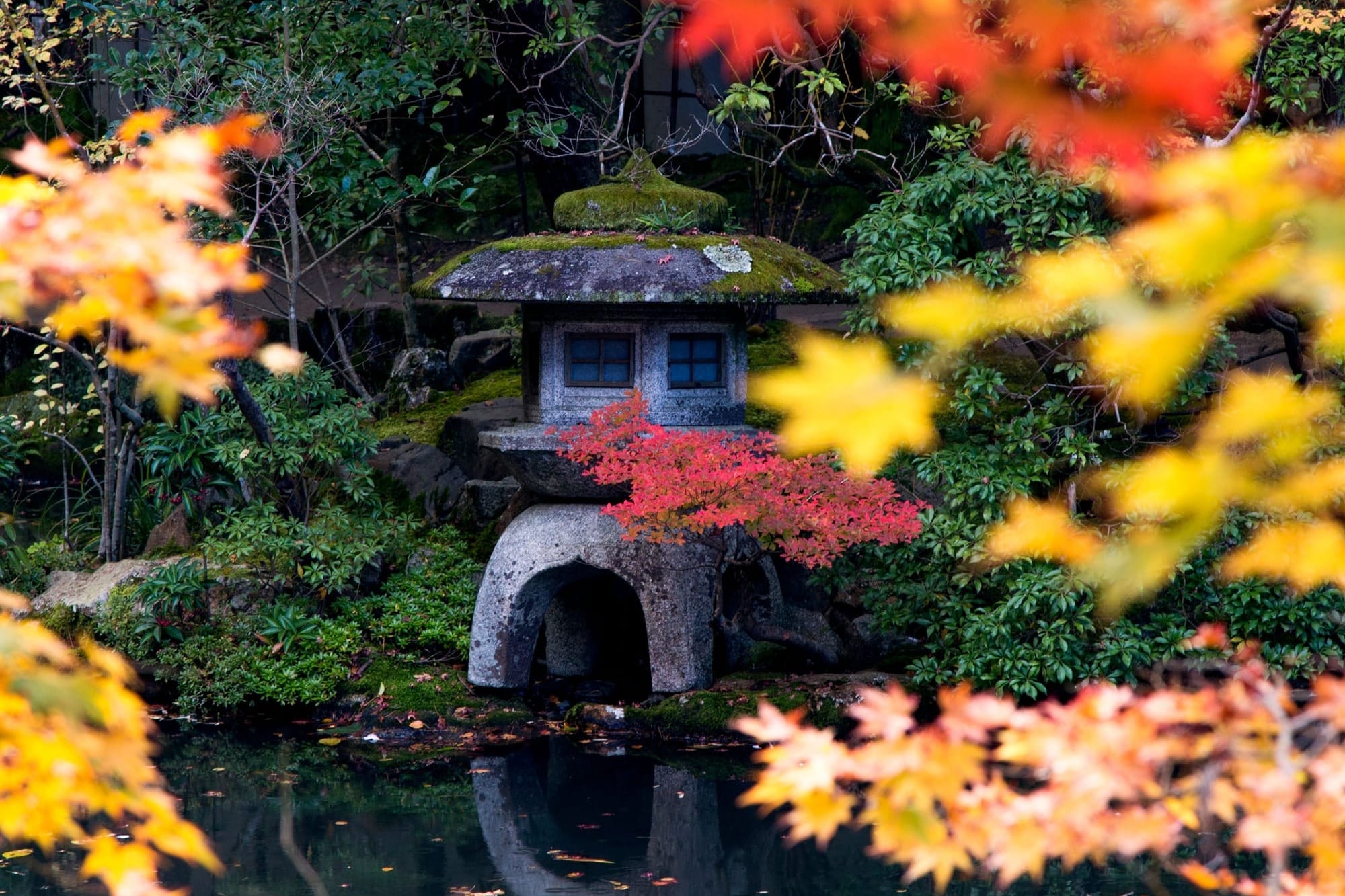 A moss-topped stone lantern reflected in a pond, framed by red and yellow autumn leaves in a Japanese garden.