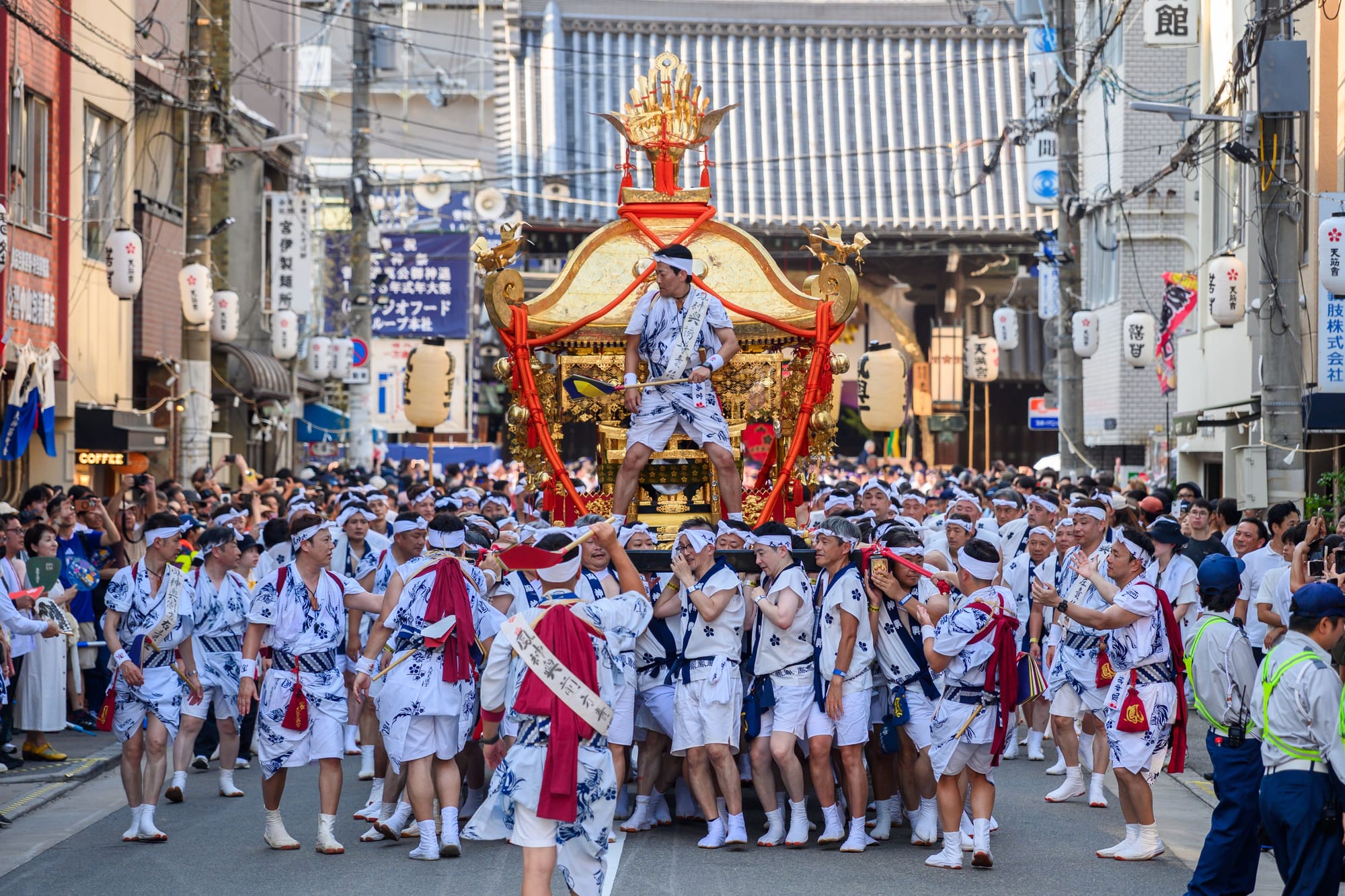 A lively Japanese festival parade fills a city street as crowds in white traditional outfits carry a richly decorated gold-and-red mikoshi, smiling.