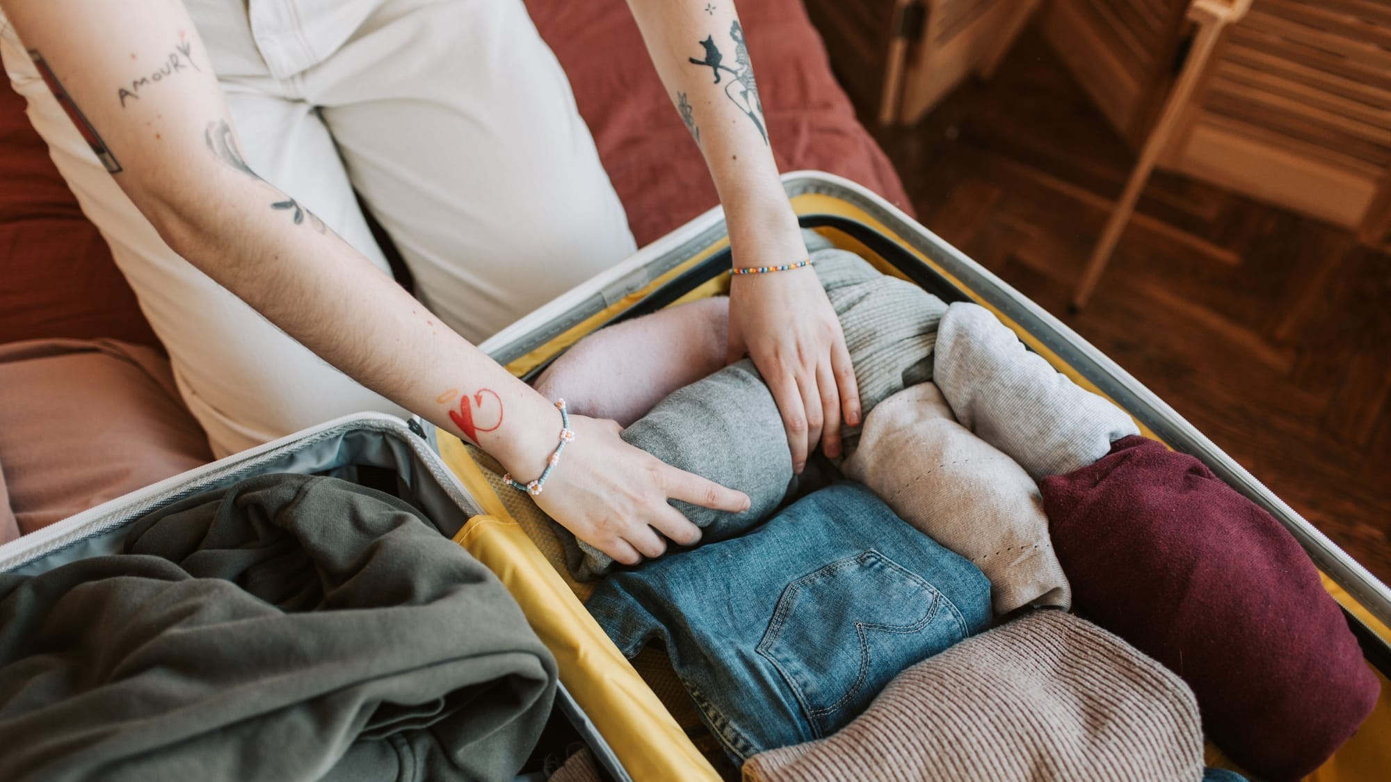 Packing for fall travel: a person with tattooed forearms presses rolled and folded clothes into an open gray suitcase with a yellow lining — blue jeans, beige corduroy, and gray and maroon knits.