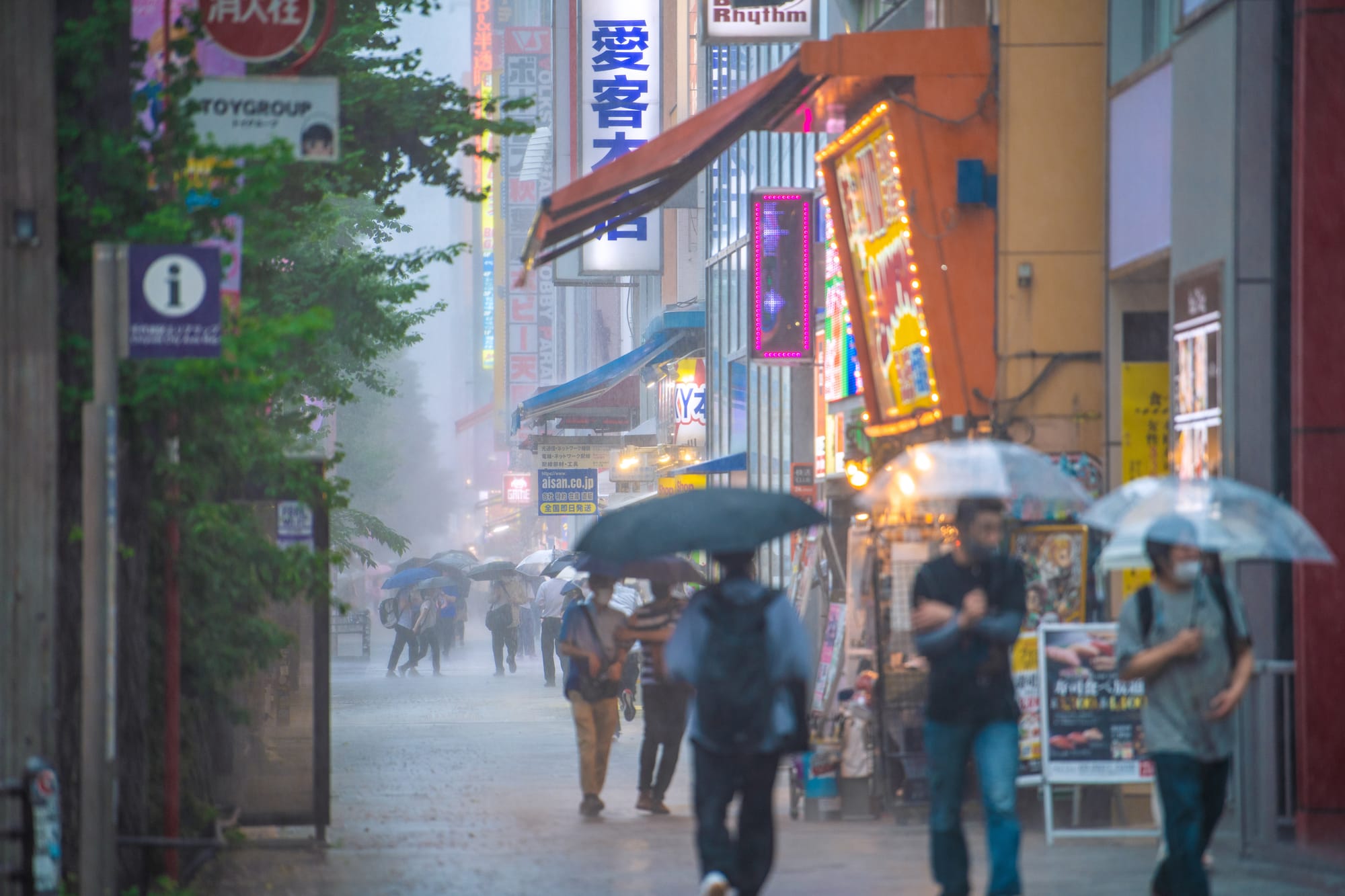 Pedestrians under clear and black umbrellas walk a wet shopping street in heavy rain, passing neon signs, hobby-shop billboards, and lit storefronts, with rain haze softening the buildings down the block.