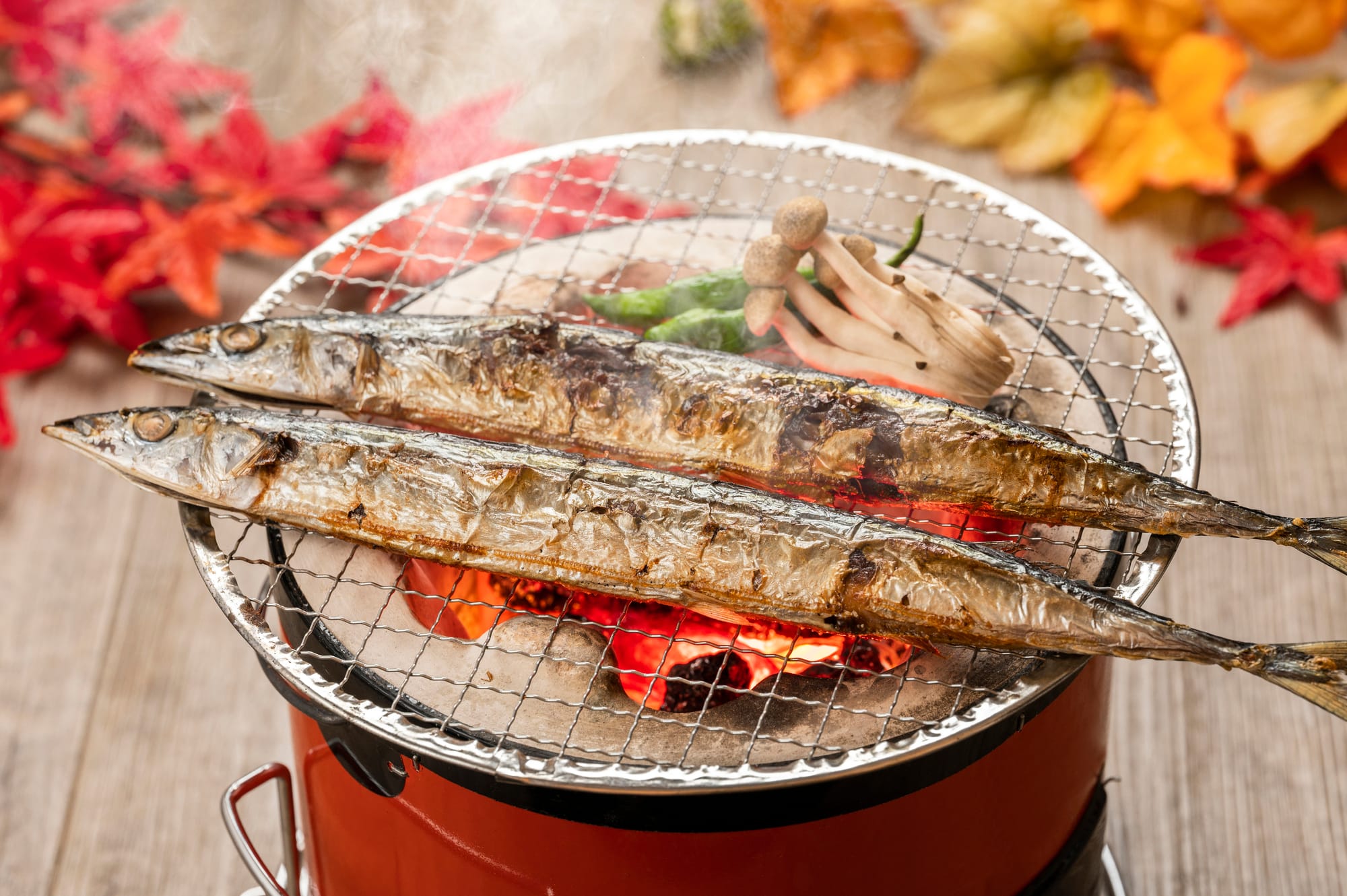 Two whole grilled fish roast on a wire rack over glowing red coals in a tabletop charcoal grill, with autumn leaves and mushrooms nearby.