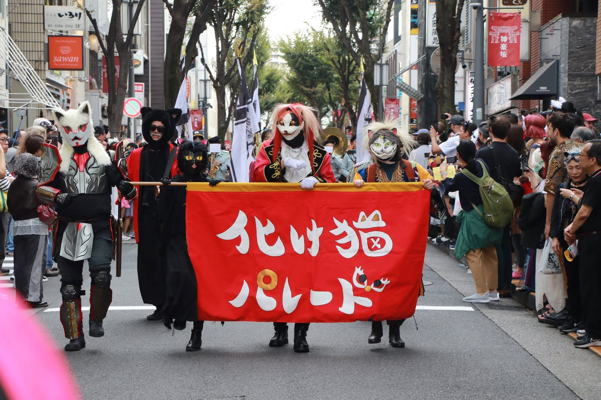 Costumed marchers in cat masks and armor carry a red banner reading 化け猫パレード (Bakeneko Parade) down a tree-lined street, flanked by spectators and red Kagurazaka lamppost banners.