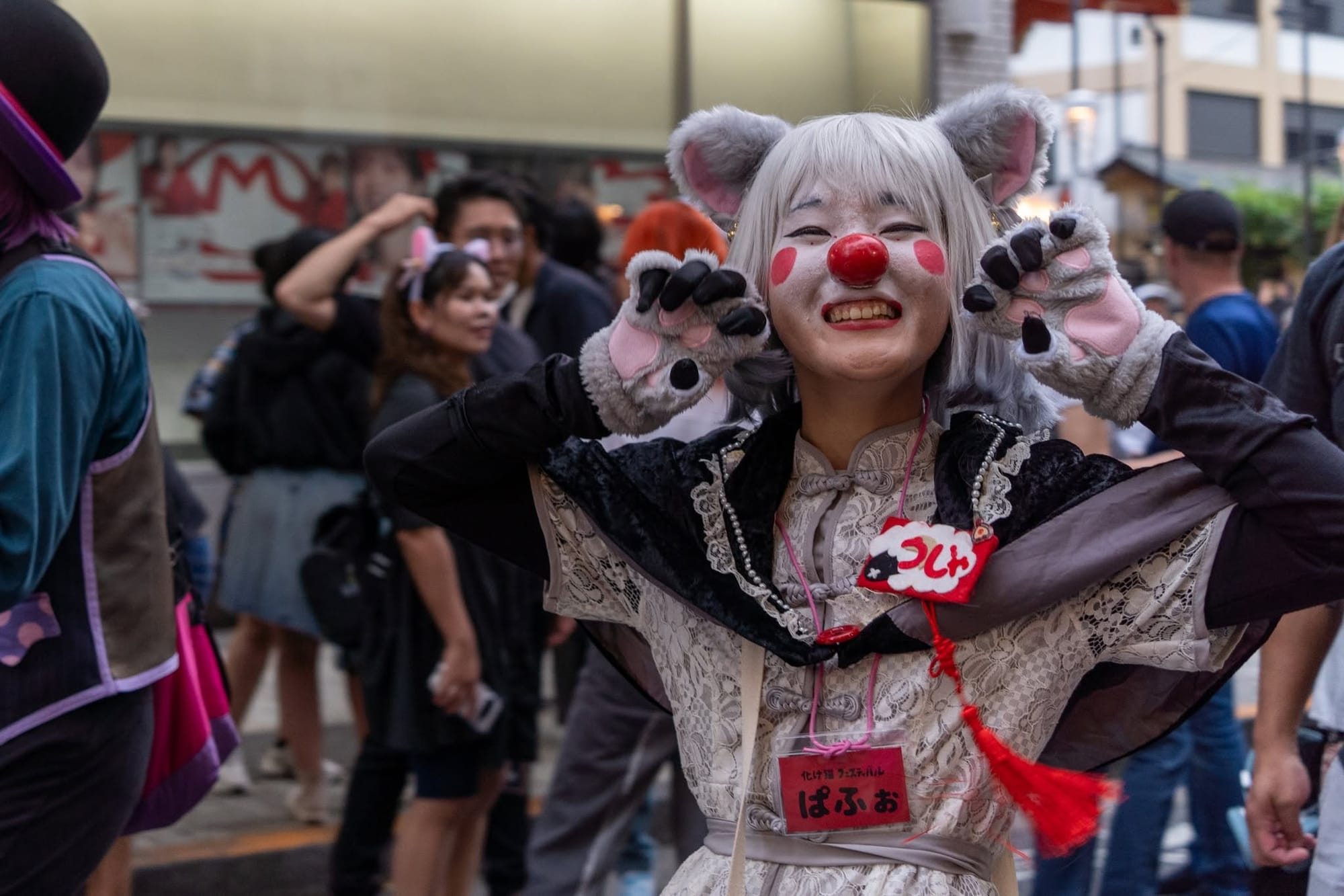 A bakeneko festival participant in a white wig, grey rounded ears, white face paint, red nose and grey paw gloves raises her claws and grins, wearing a 化け猫フェスティバル name badge over a cream lace dress.