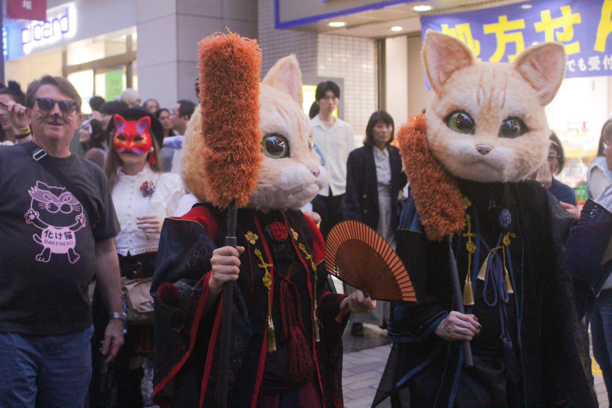 Two marchers in oversized plush cat heads with large green eyes and dark tasseled robes hold a folding fan and fluffy orange staffs, while a spectator in a 化け猫 BAKENEKO T-shirt looks on.