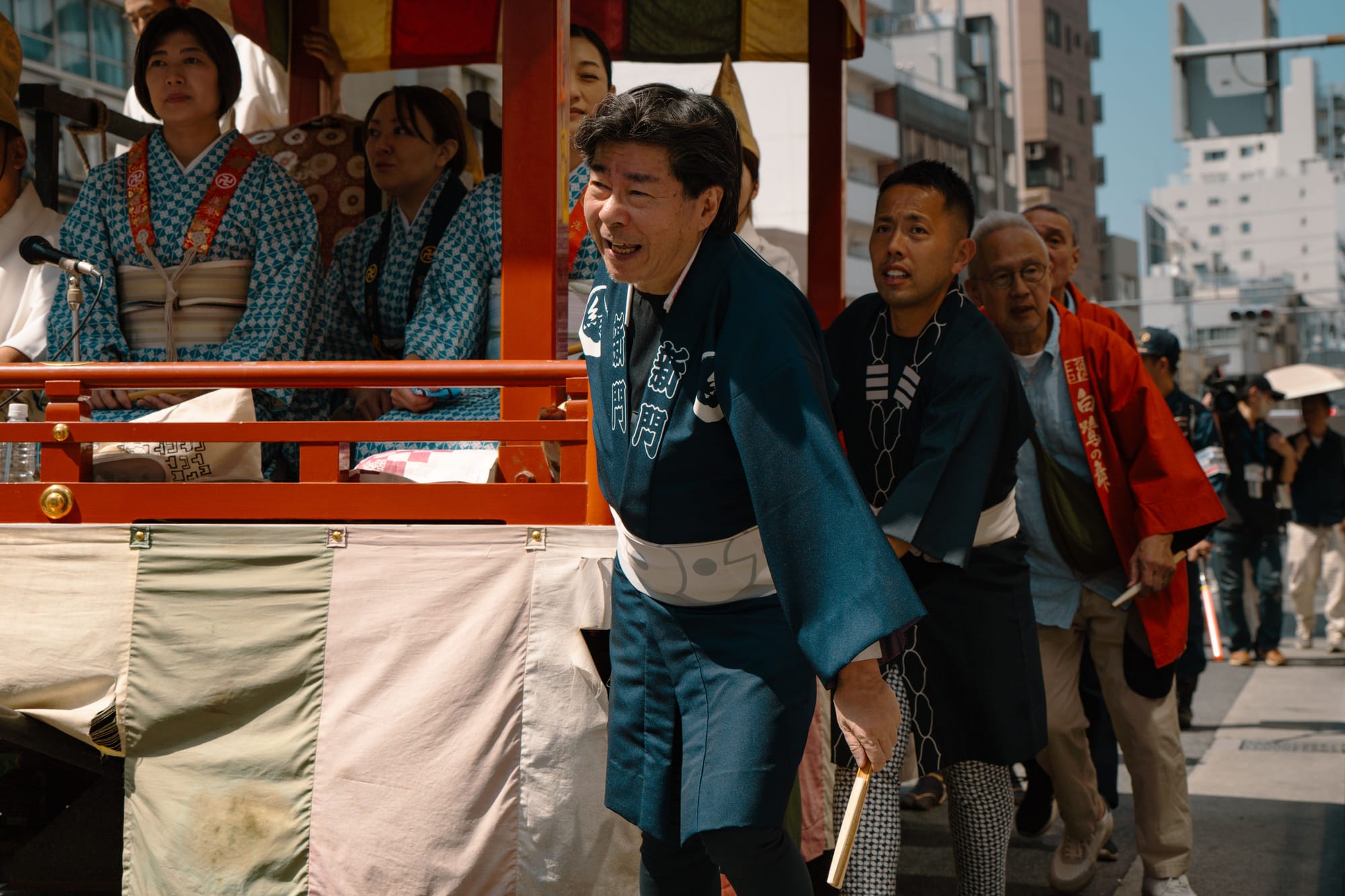 Men in indigo happi coats pulling a red festival float past modern city buildings.