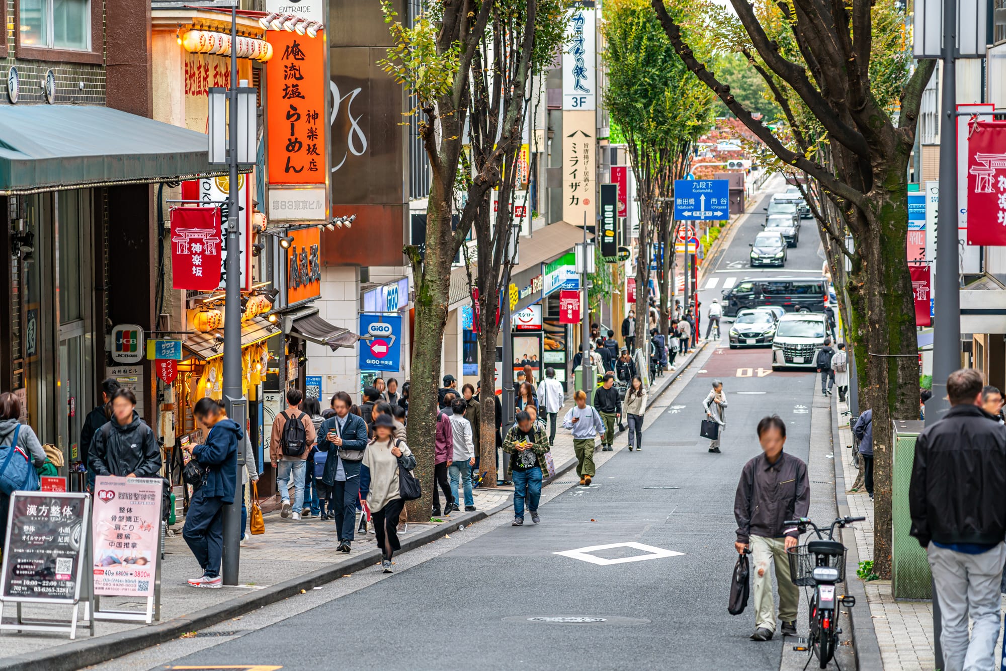 A sloping Kagurazaka shopping street lined with trees, red 神楽坂 banners and signs for ramen shops, an arcade and restaurants, with pedestrians on the sidewalk and cars parked along the road.