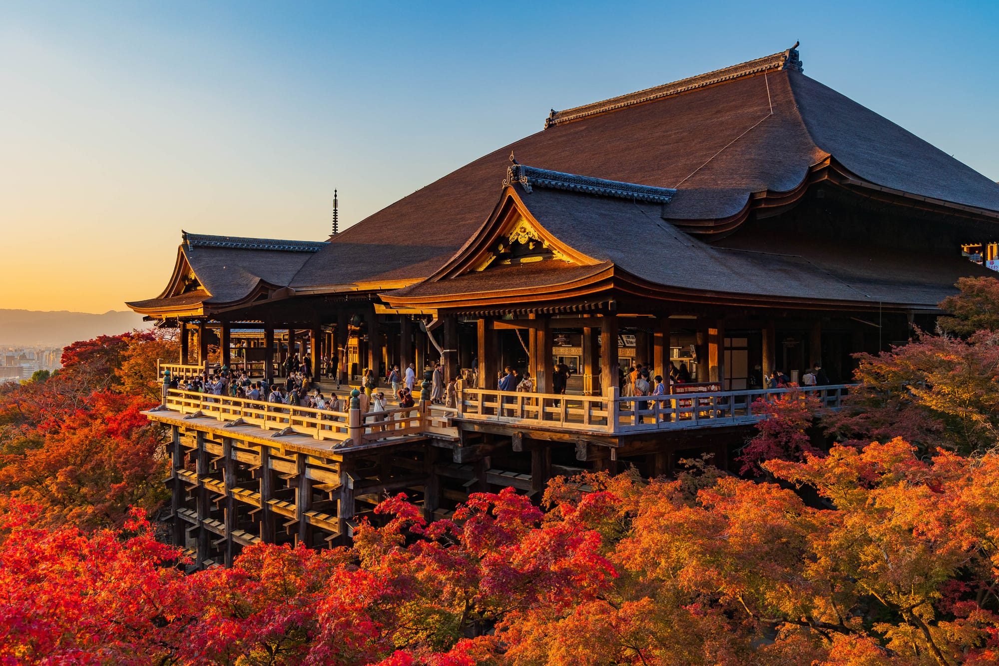 Visitors crowd the wooden veranda of a large temple hall raised on tall timber pillars, surrounded by red and orange maple fall foliage, with hills and a city beyond at sunset.