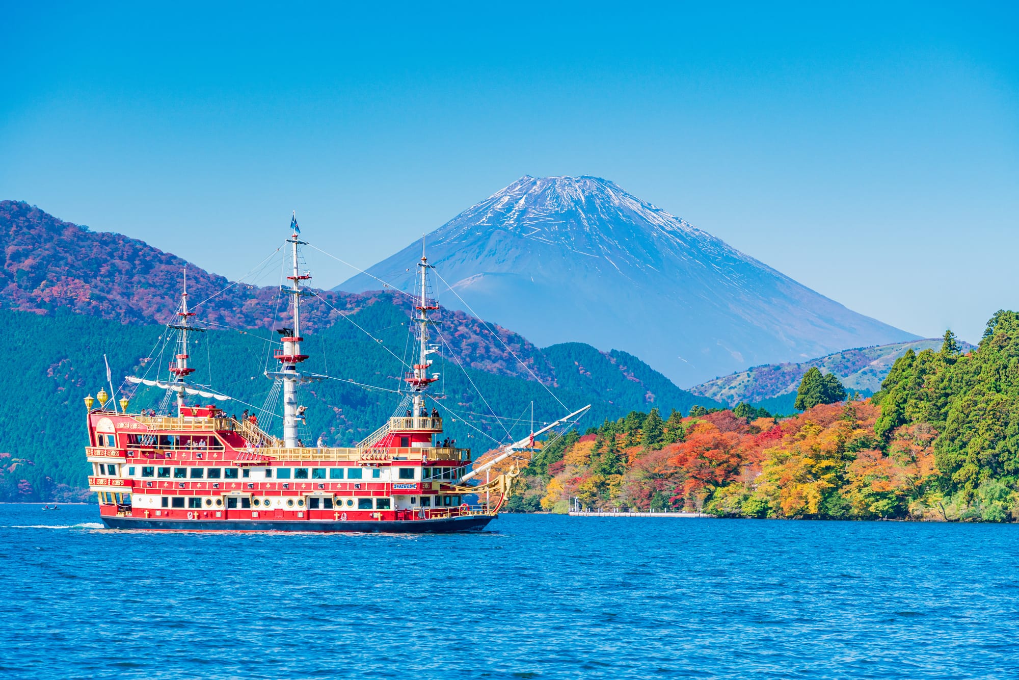 A red and white pirate-style sightseeing ship crosses a deep blue lake below snow-capped Mount Fuji, with hillsides of red, orange, and evergreen fall foliage rising along the far shore under clear sky.