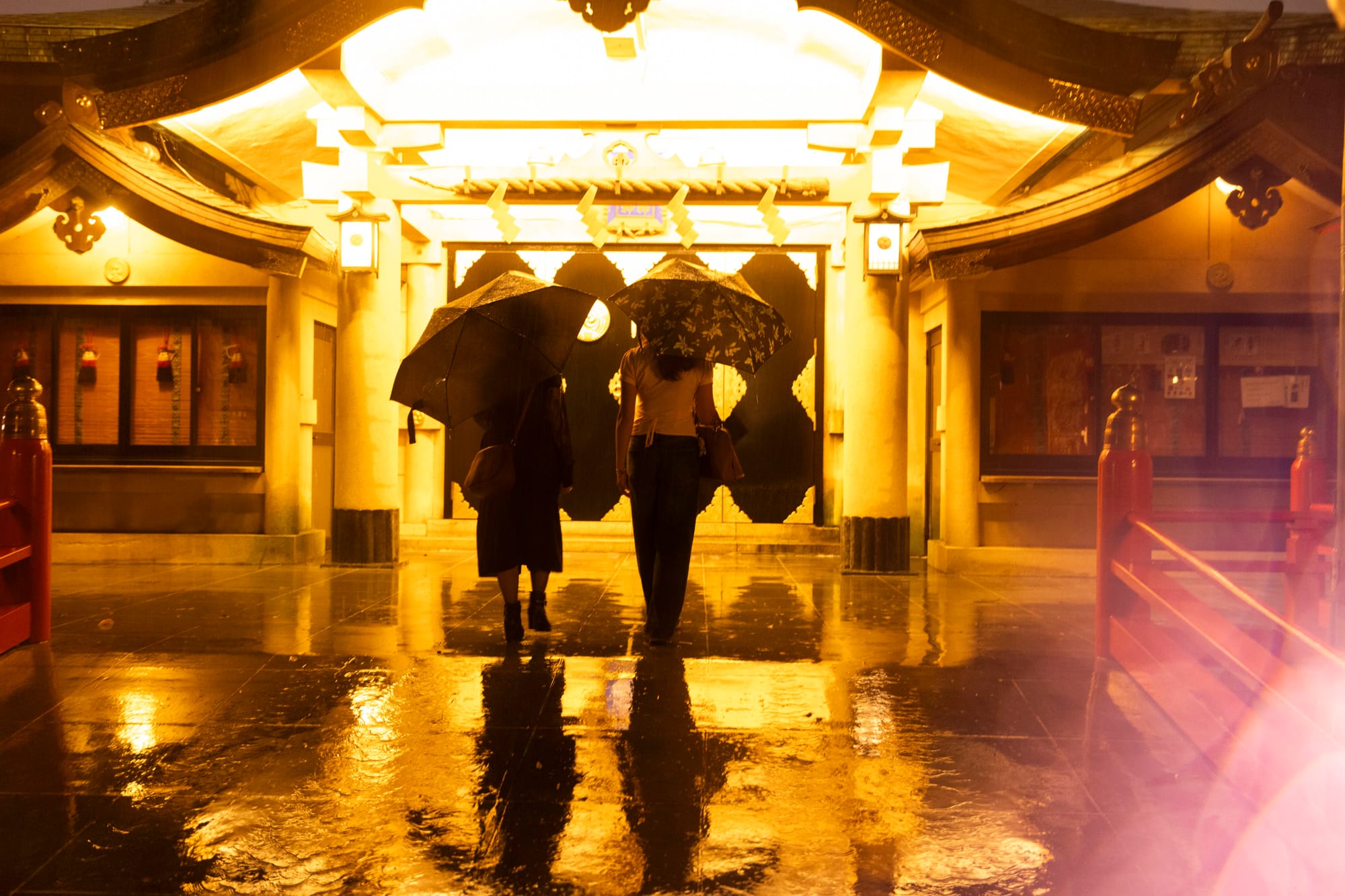 Two people with umbrellas approach a lit shrine gate across rain-soaked stone at night