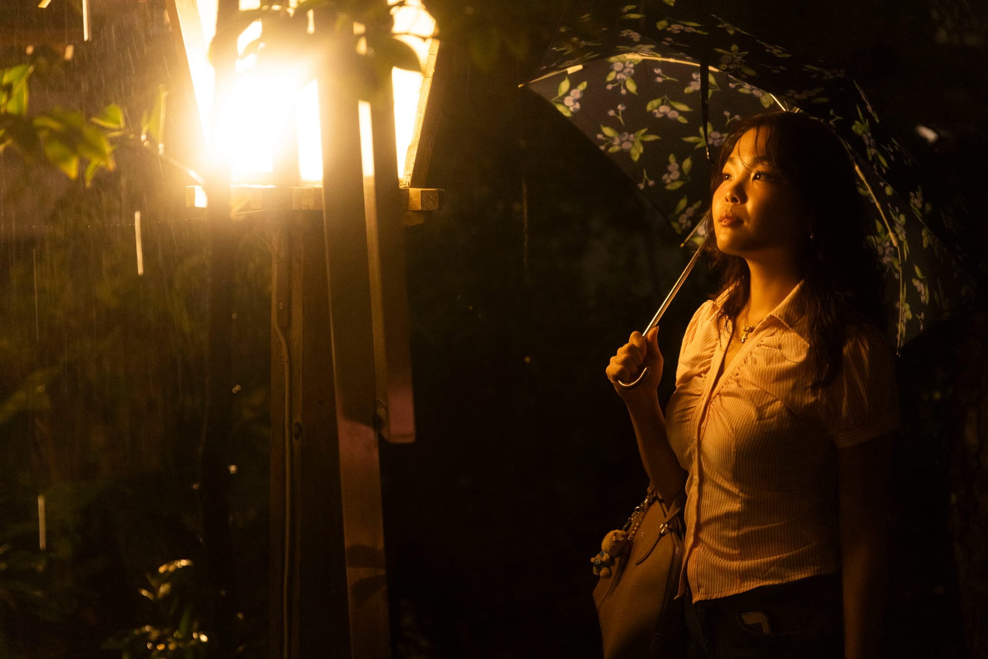 Woman with a floral umbrella looks up at a glowing wooden lantern in the rain at night