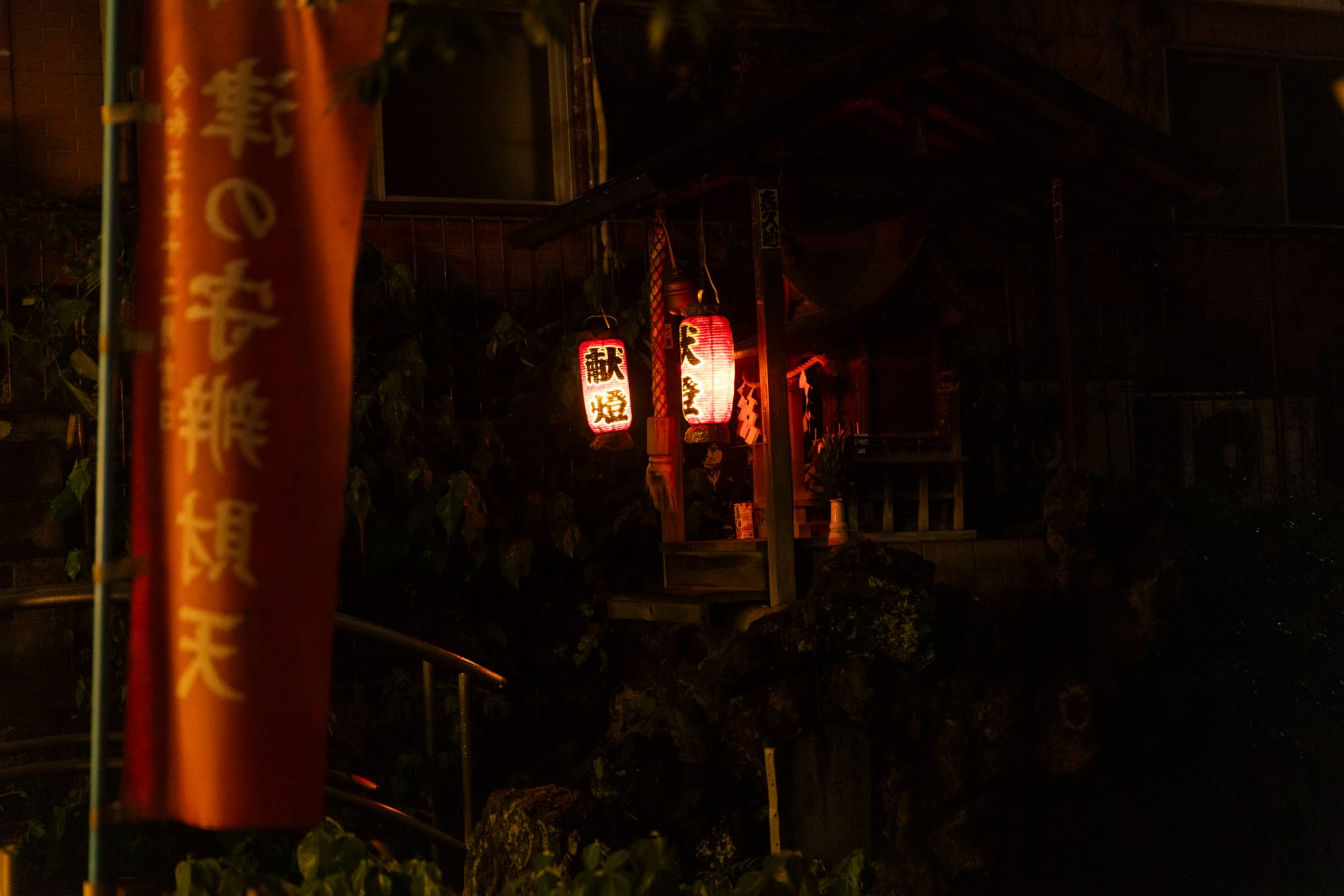 Two glowing red paper lanterns hang at a small shrine at night, with a red Tsu-no-Kami Benzaiten banner blurred in the foreground