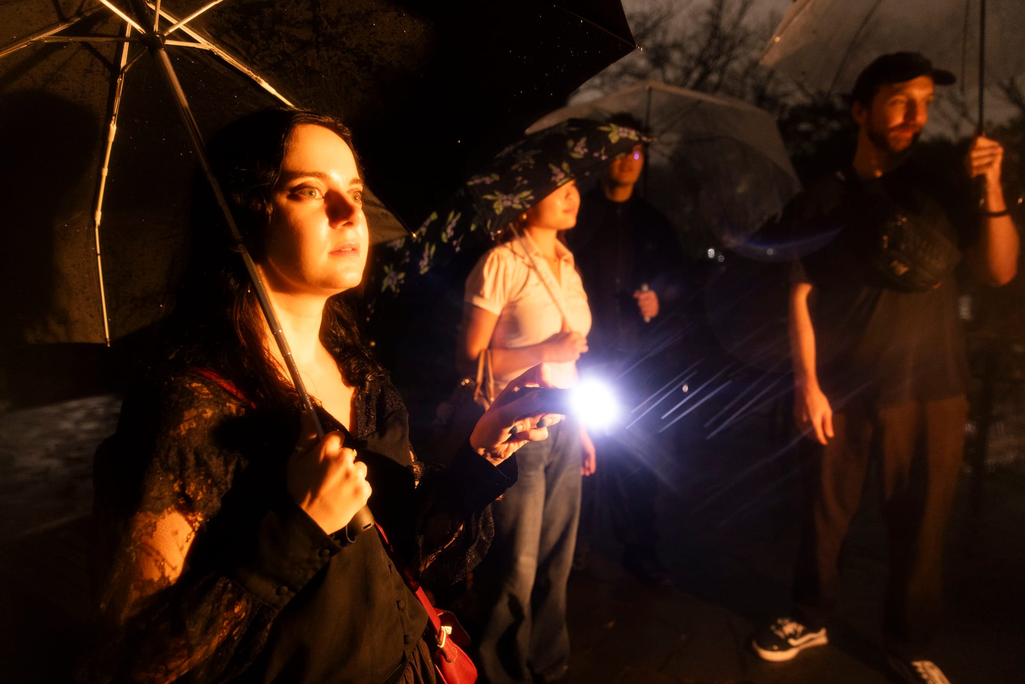Woman holding a flashlight and umbrella at night, with a small group behind her in the rain