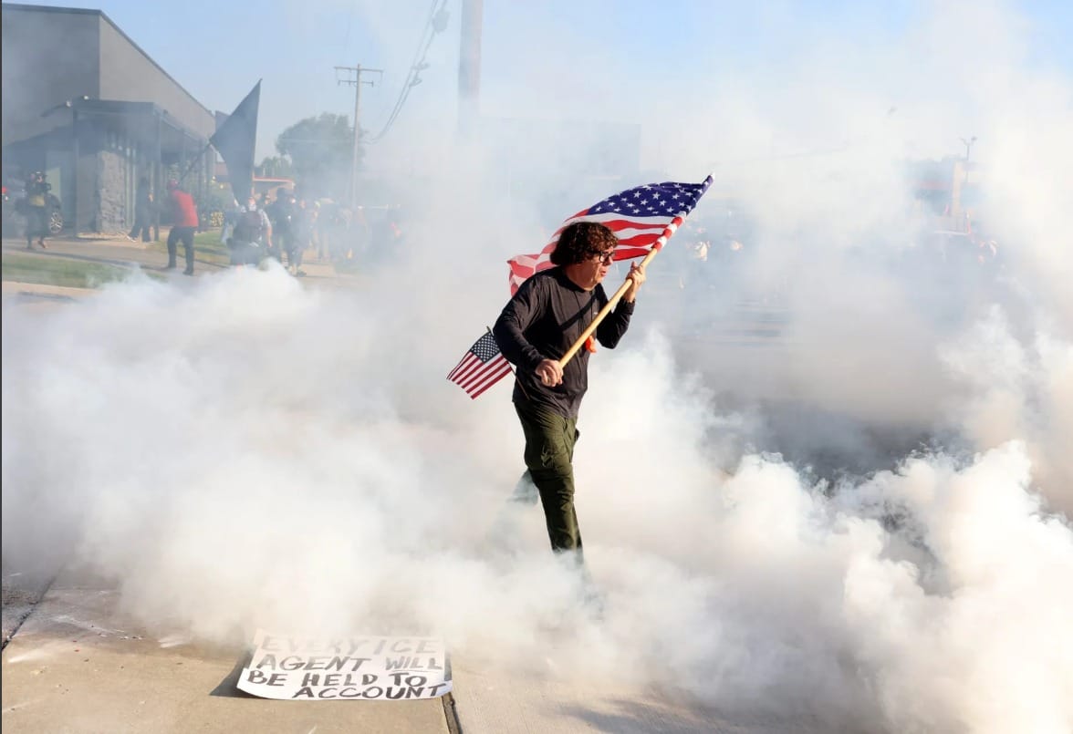 Marine veteran Curtis Evans, with a backup sidearm flag (two is one and one is none!), being gassed by Trump goons at an Illinois ICE facility on September 19, 2025. A sign on the ground says "EVERY ICE AGENT WILL BE HELD TO ACCOUNT". 