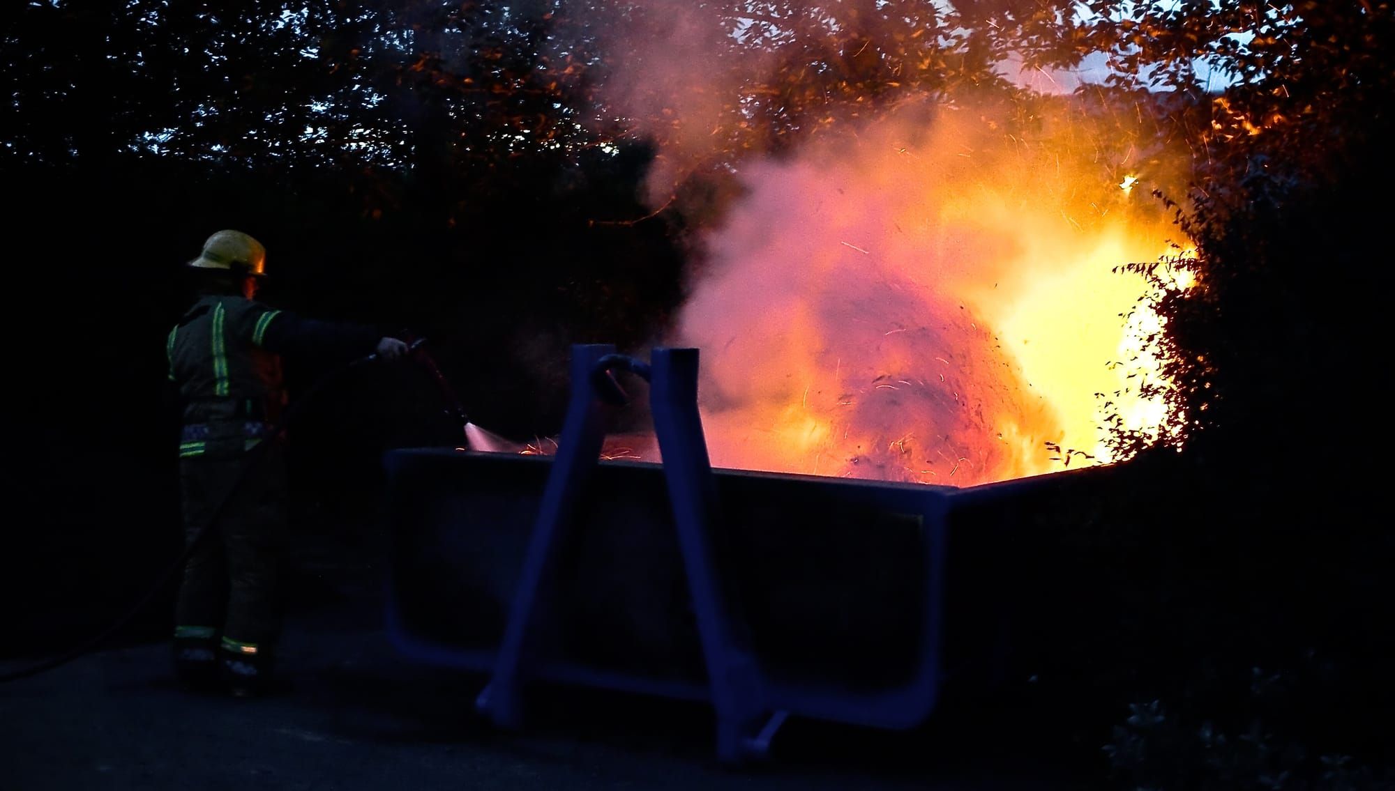 Photo of a dumpster on fire with a firefighter in the left foreground, and trees in the background
