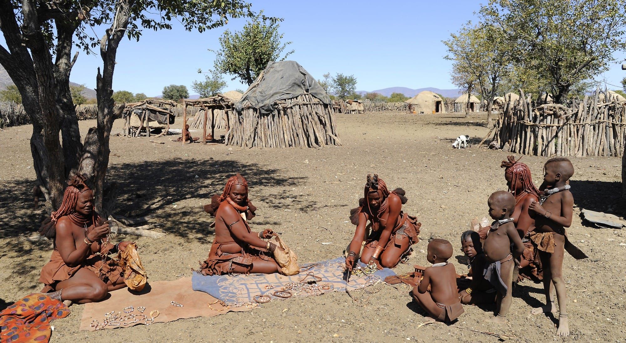 Three Himba women sit on blankets with beadwork while five children look on; in the background are round huts, a round livestock pen, and trees
