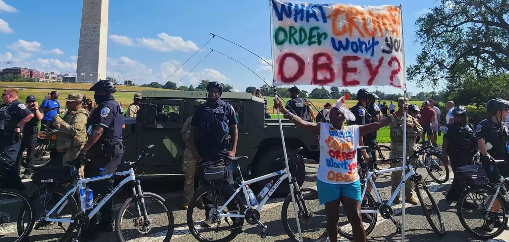 With Washington Monument in the background, federalized cops and militarized vehicles are staged with the Washington Monument in the background as a middle aged Black woman protests with a c
